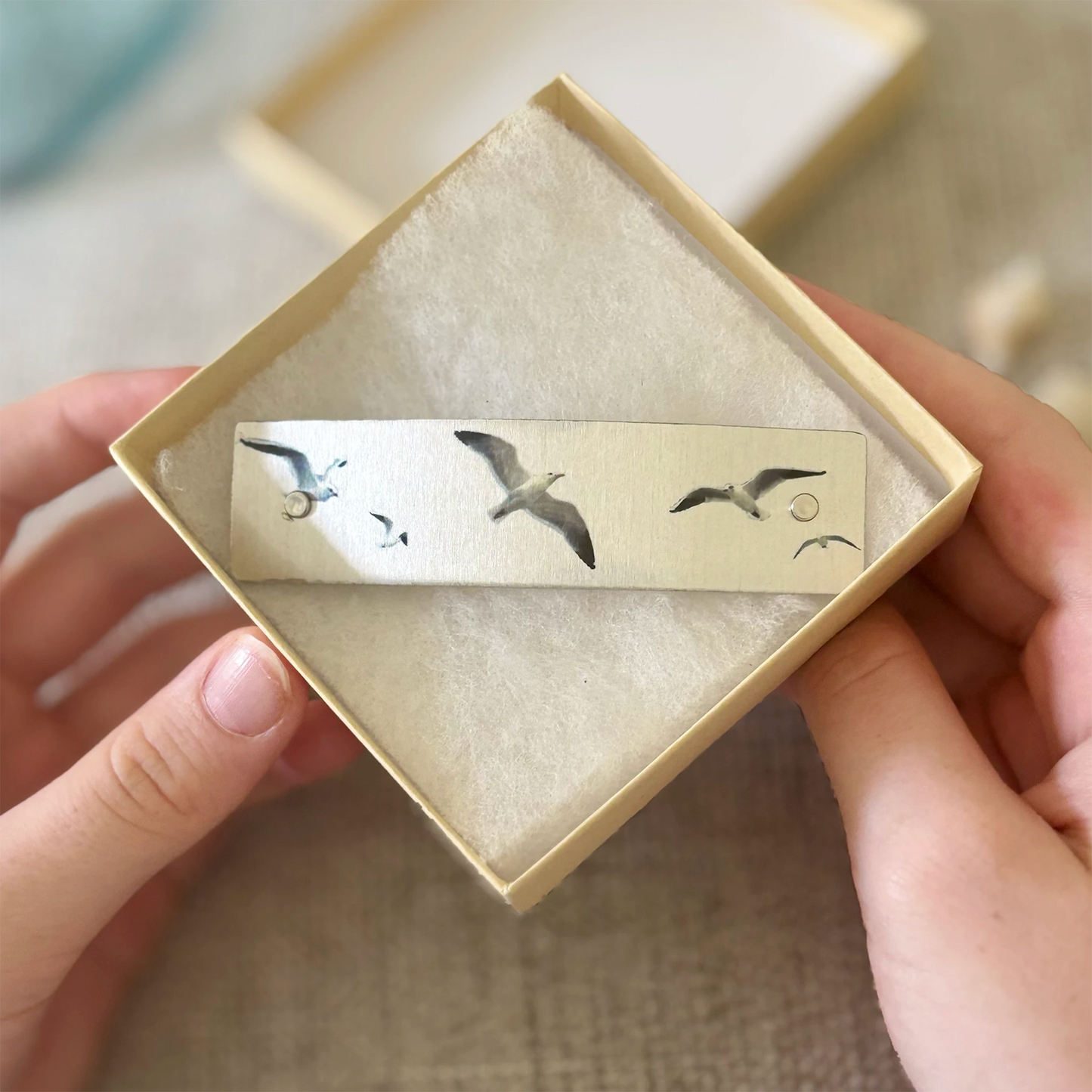 A close-up of hands holding a small square cream-colored jewelry box containing a silver-toned barrette decorated with flying seagulls.