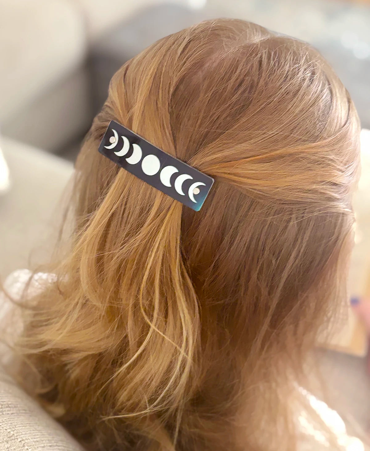 A close-up lifestyle shot of a person's blonde hair secured with a black hair clip decorated with silver moon phases, set against a soft-focus indoor background.