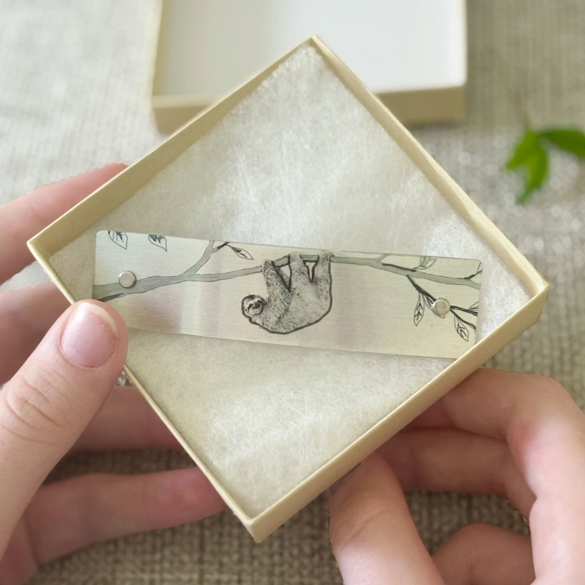 A silver-toned barrette with a black line art sketch of a sloth hanging from a branch, presented inside a square cream-colored gift box held by hands.