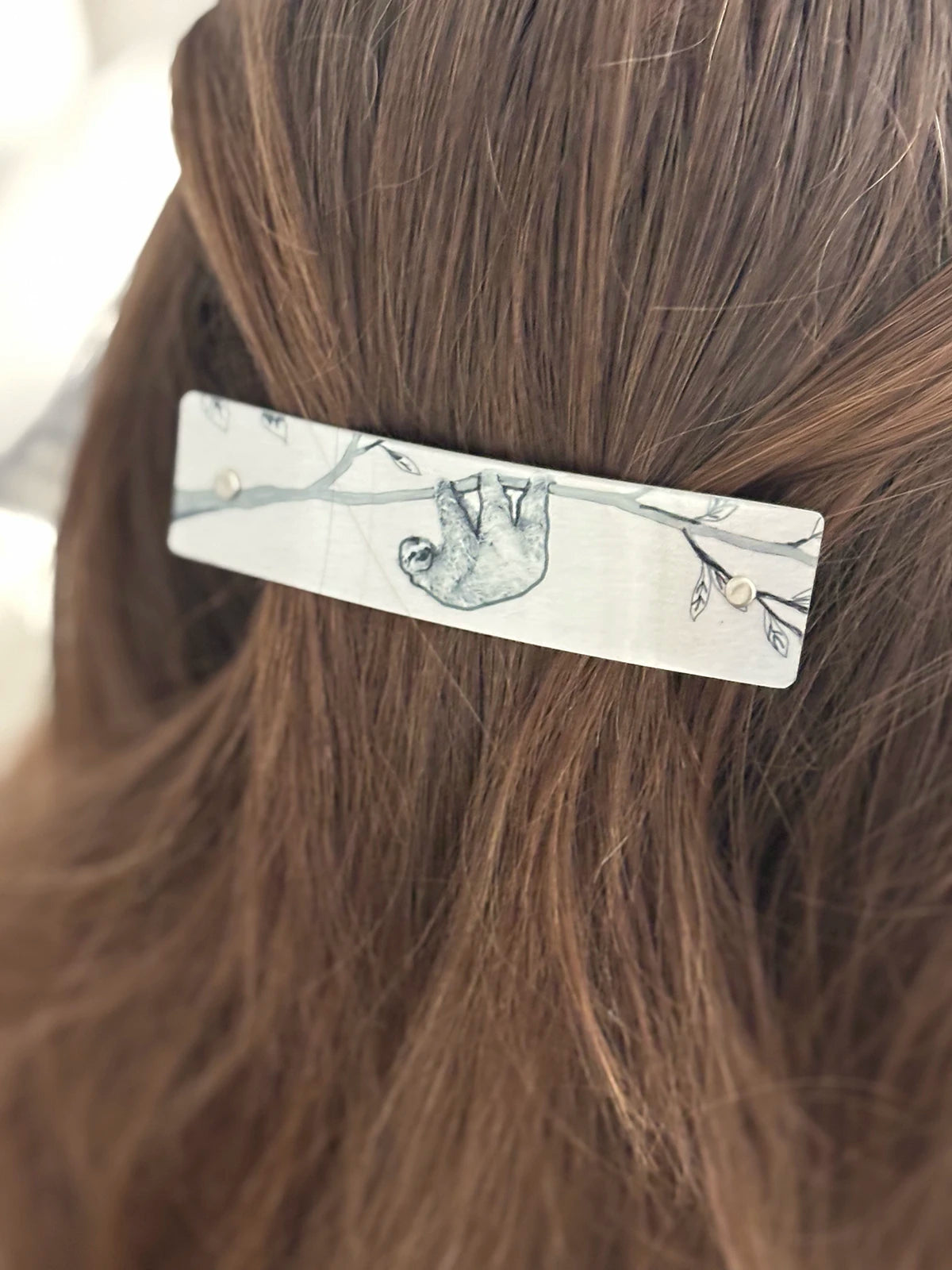 A close-up of a boutique hair clip with a silver background with a black line-art illustration of a sloth hanging from a branch.