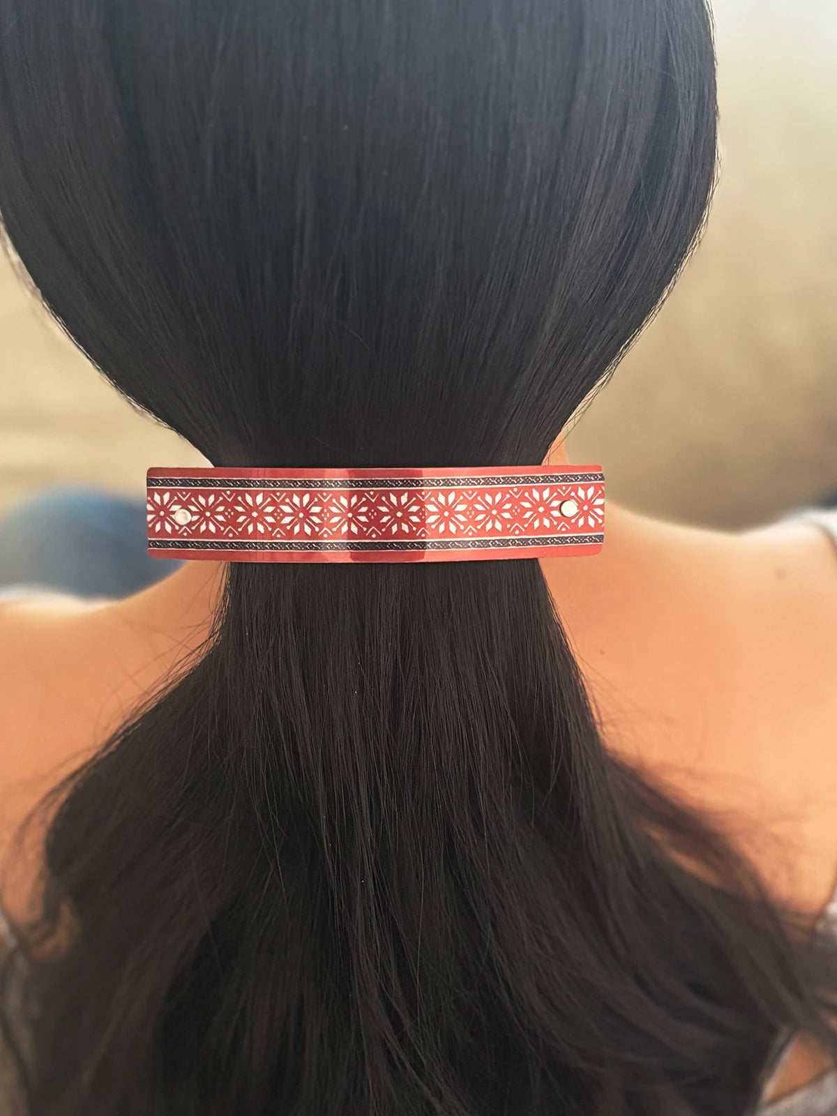 A close-up of a person's dark hair tied back with a slim metallic hair barrette featuring a red and silver geometric pattern.