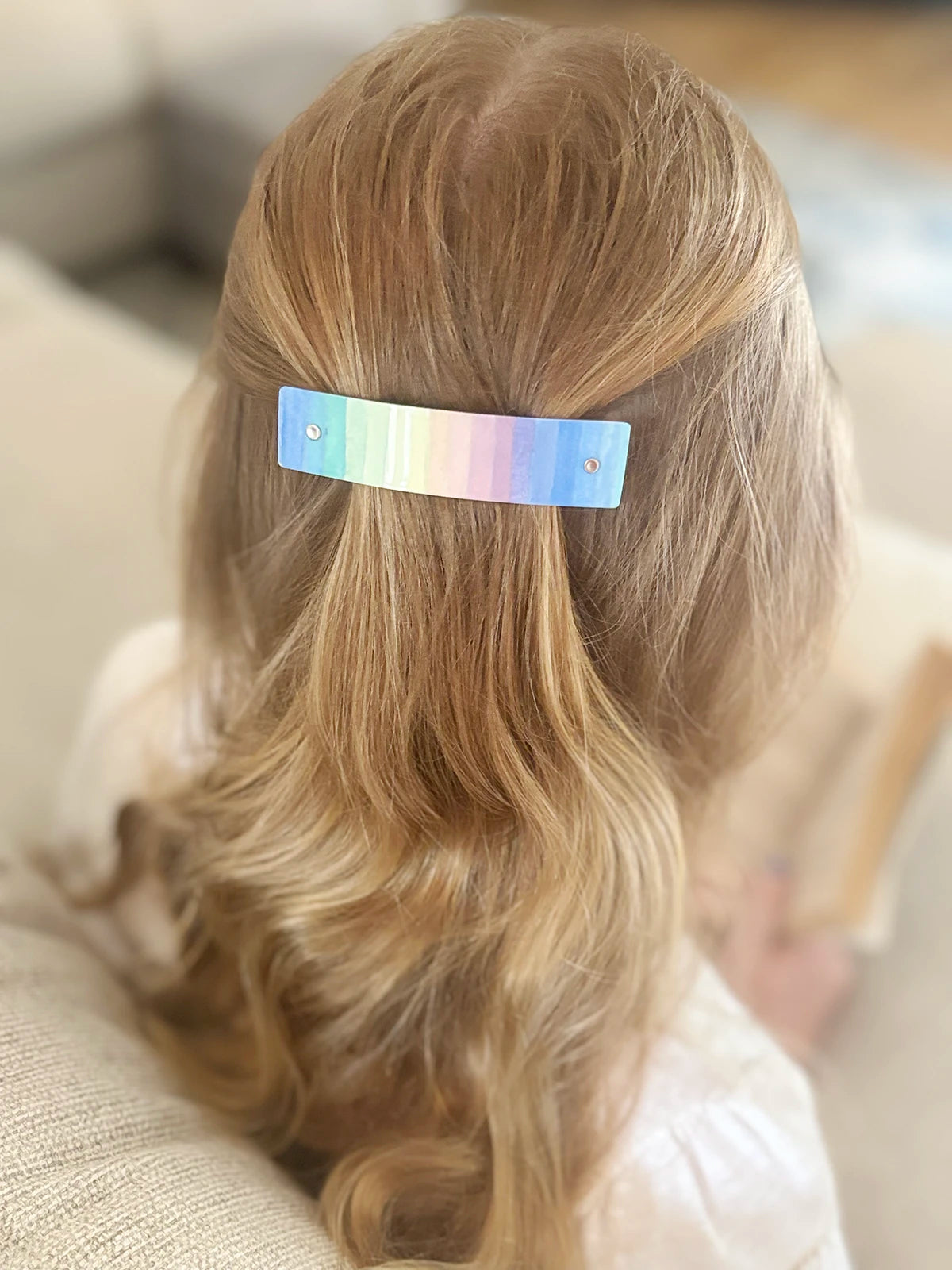 A close-up back view of a person with long blonde wavy hair wearing a slim rectangular hair clip with a pastel rainbow finish.