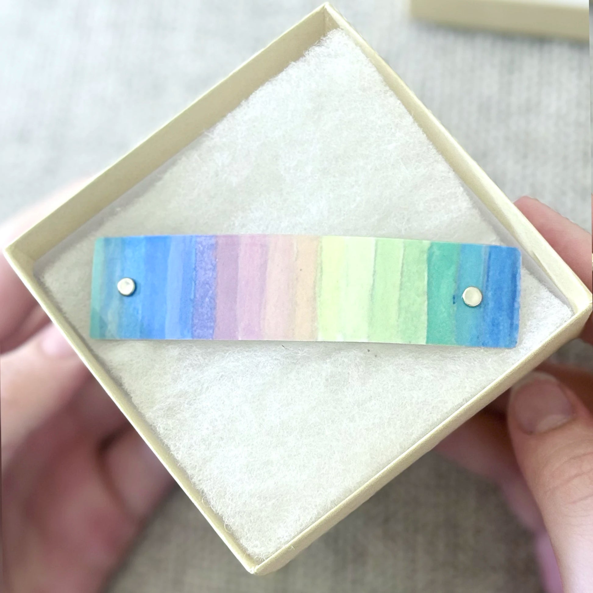 A rectangular pastel rainbow striped barrette with two small silver metal rivets, presented inside a cream-colored square gift box.. Hands are holding the box.