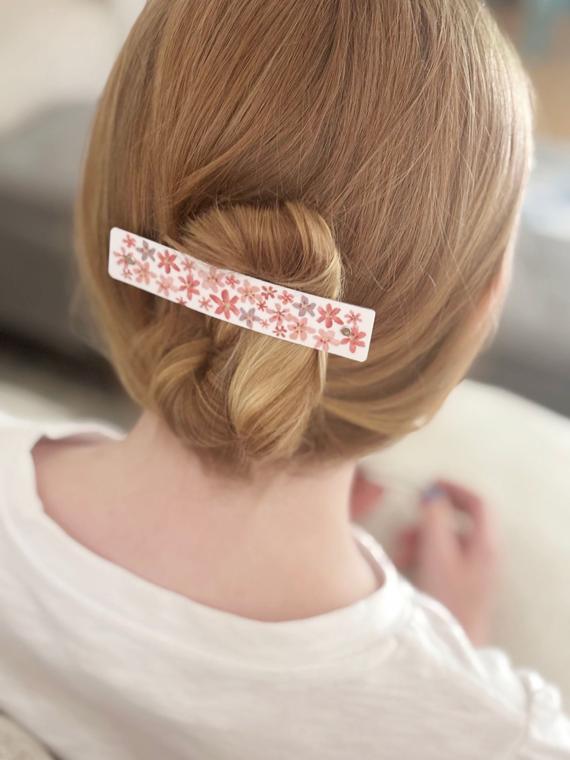 A close-up back view of a person with light brown hair styled in a low bun, secured with a white hair clip decorated with a pink and coral  floral pattern.