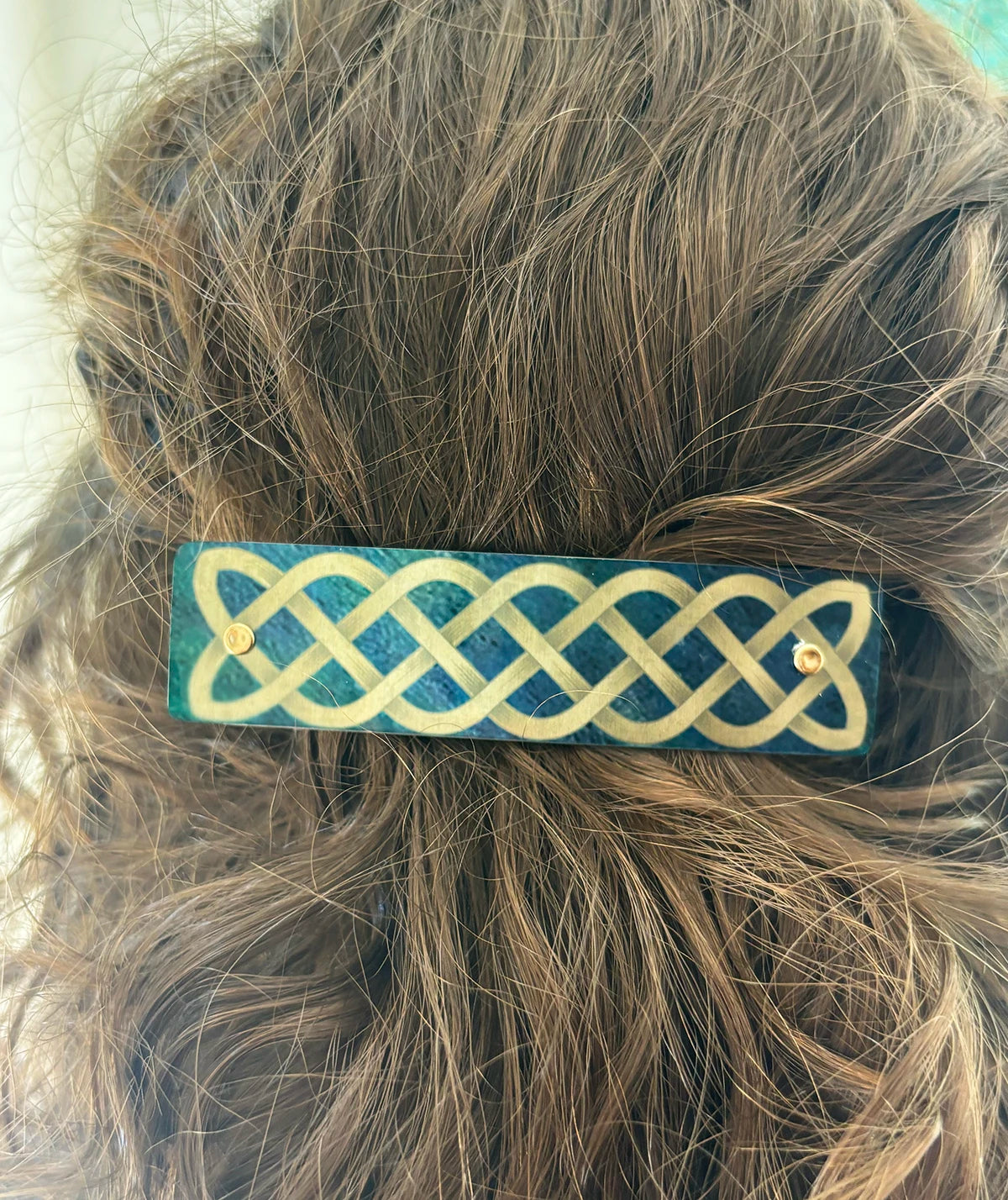A close-up of brown hair secured with a rectangular hair clip featuring a green background and gold-colored Celtic knot pattern.