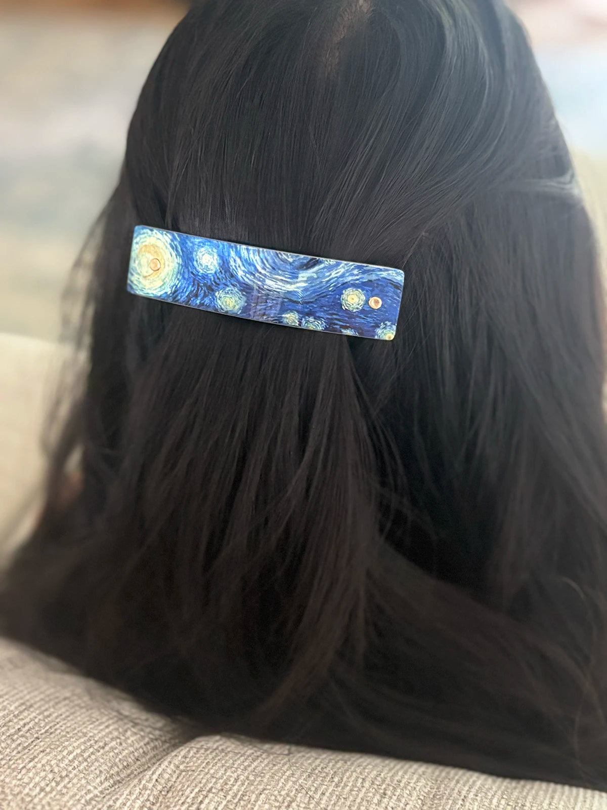 A close-up of a person's dark hair from behind, featuring a rectangular hair clip decorated with a blue and yellow Starry Night painting pattern.