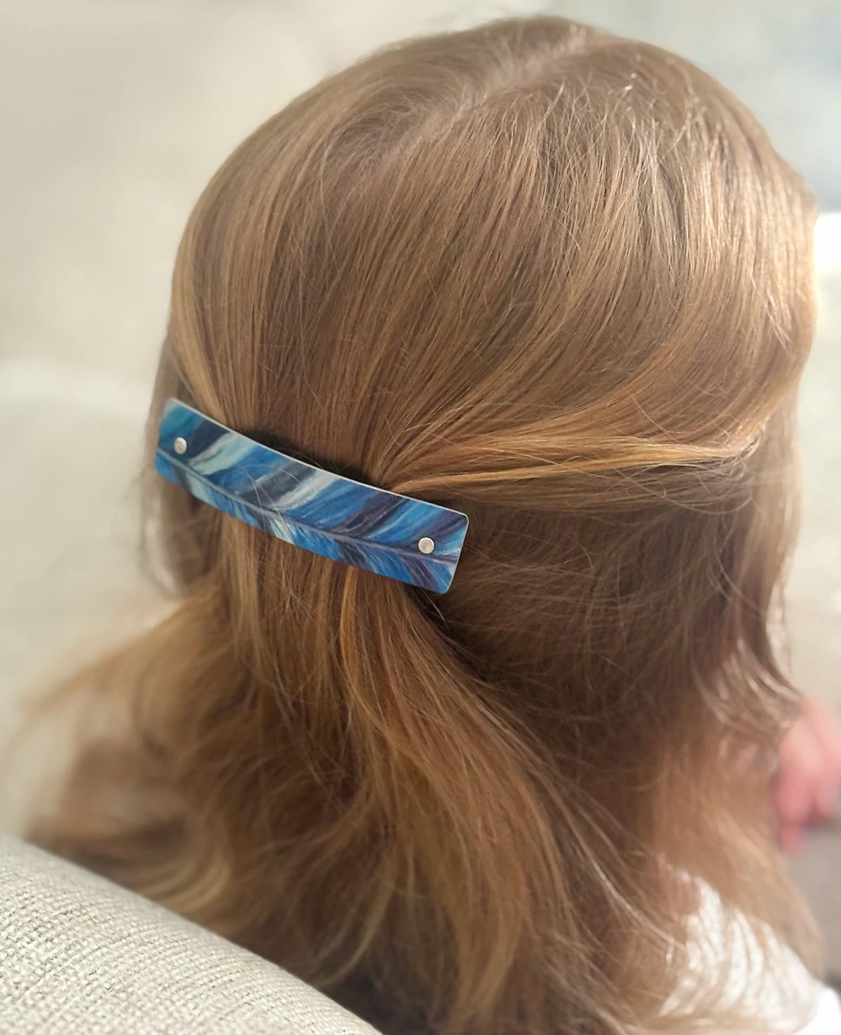 A close-up shot from behind of a person with strawberry blond hair wearing a blue and white feather-patterned hair clip.