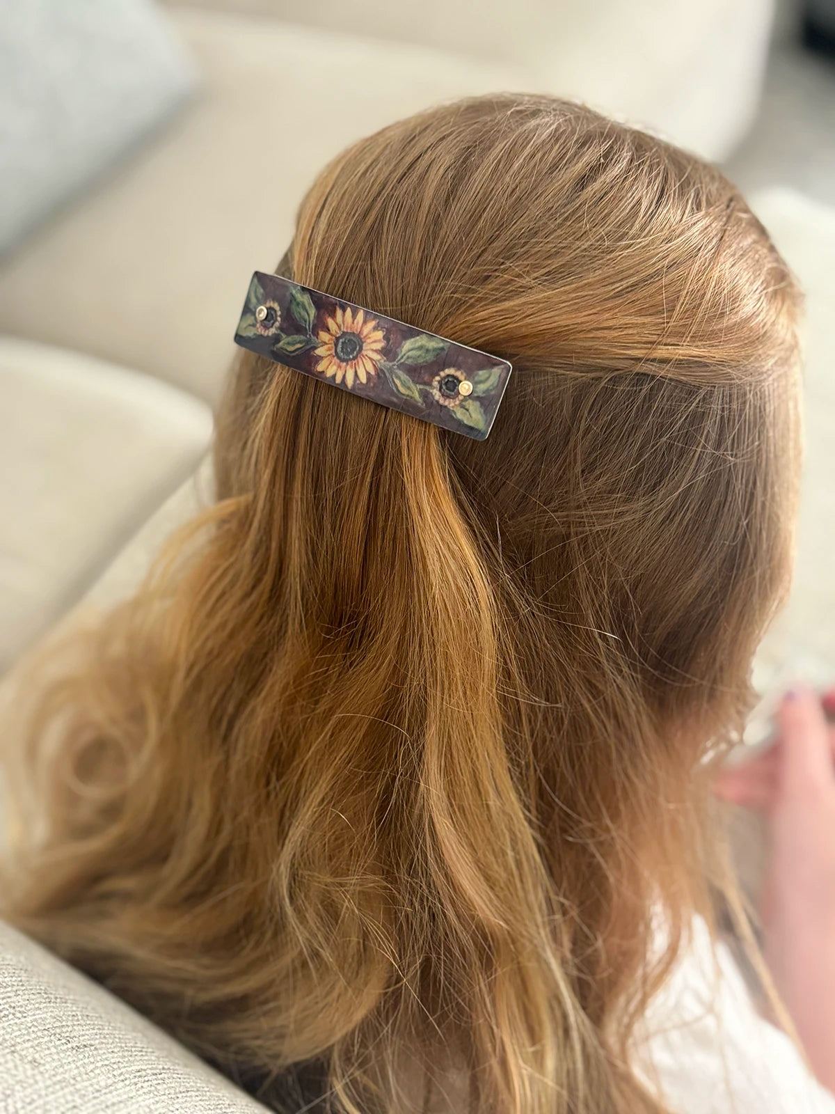 A close-up lifestyle shot of a person with long blonde hair wearing a decorative hair clip. The rectangular brown hair clip features a hand-painted floral design with a prominent sunflower in the center and green leaves.