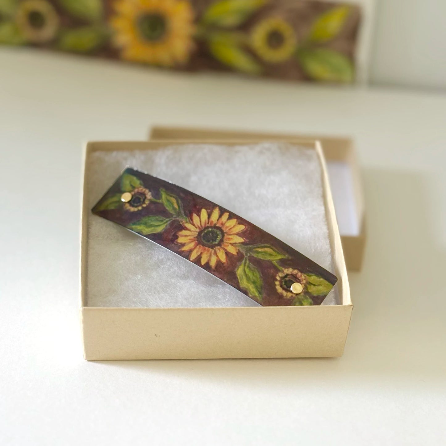 A rectangular hair clip featuring a sunflower and green leaf pattern on a dark background, displayed inside a small Kraft paper gift box.