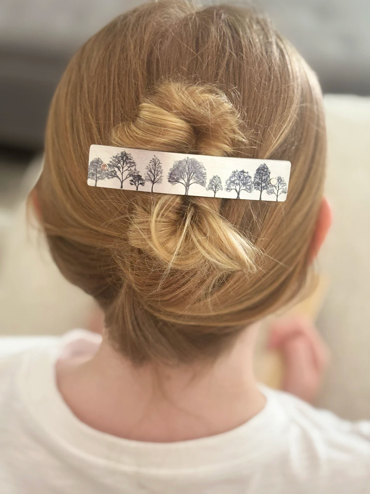 A close-up back view of a person with blonde hair styled in a low bun, secured with a slim handmade hair clip decorated with a black tree silhouette pattern on a silver background.