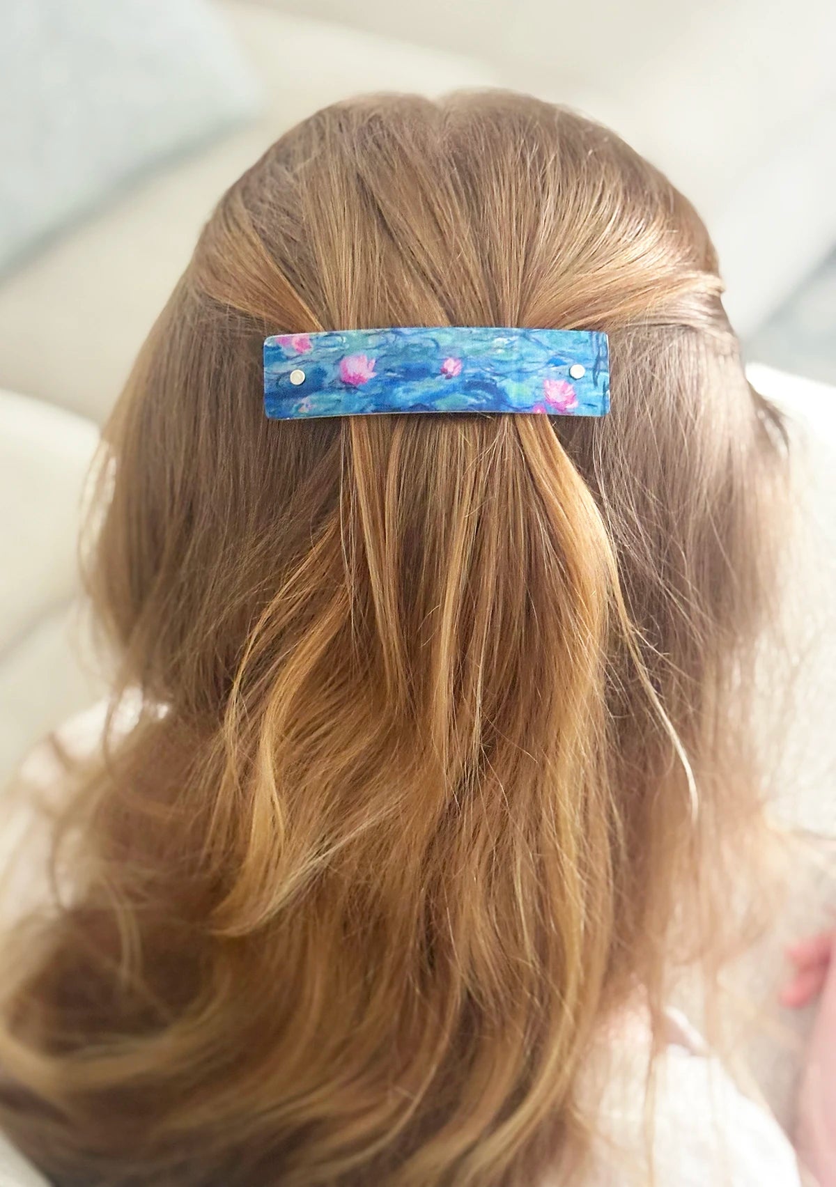 A close-up back view of a person with long blond hair wearing a rectangular hair clip. The clip has a blue background with a painted floral design of water lines.