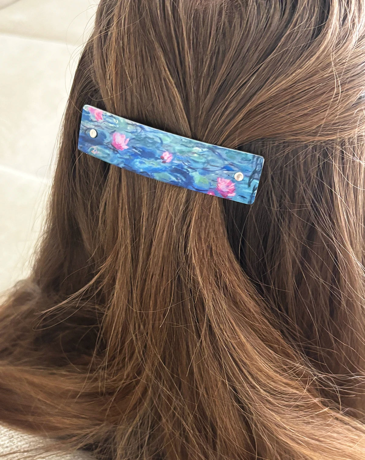 A close-up of a person's brown hair secured with a rectangular hair clip featuring a blue and pink water lily painting design.