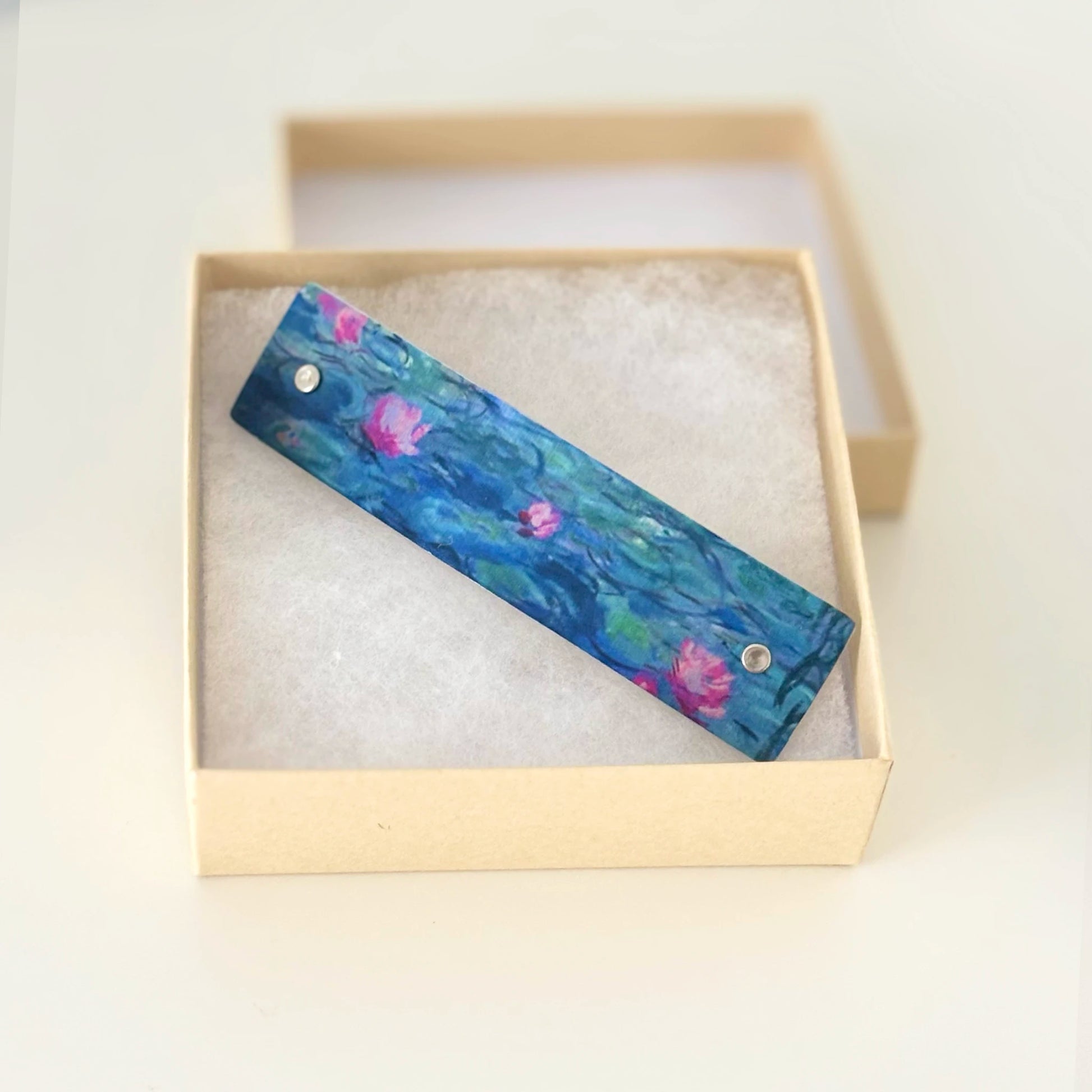 A rectangular hair barrettet featuring a painted water lily design in shades of blue, green, and pink, displayed inside an open beige gift box with white foam insert.