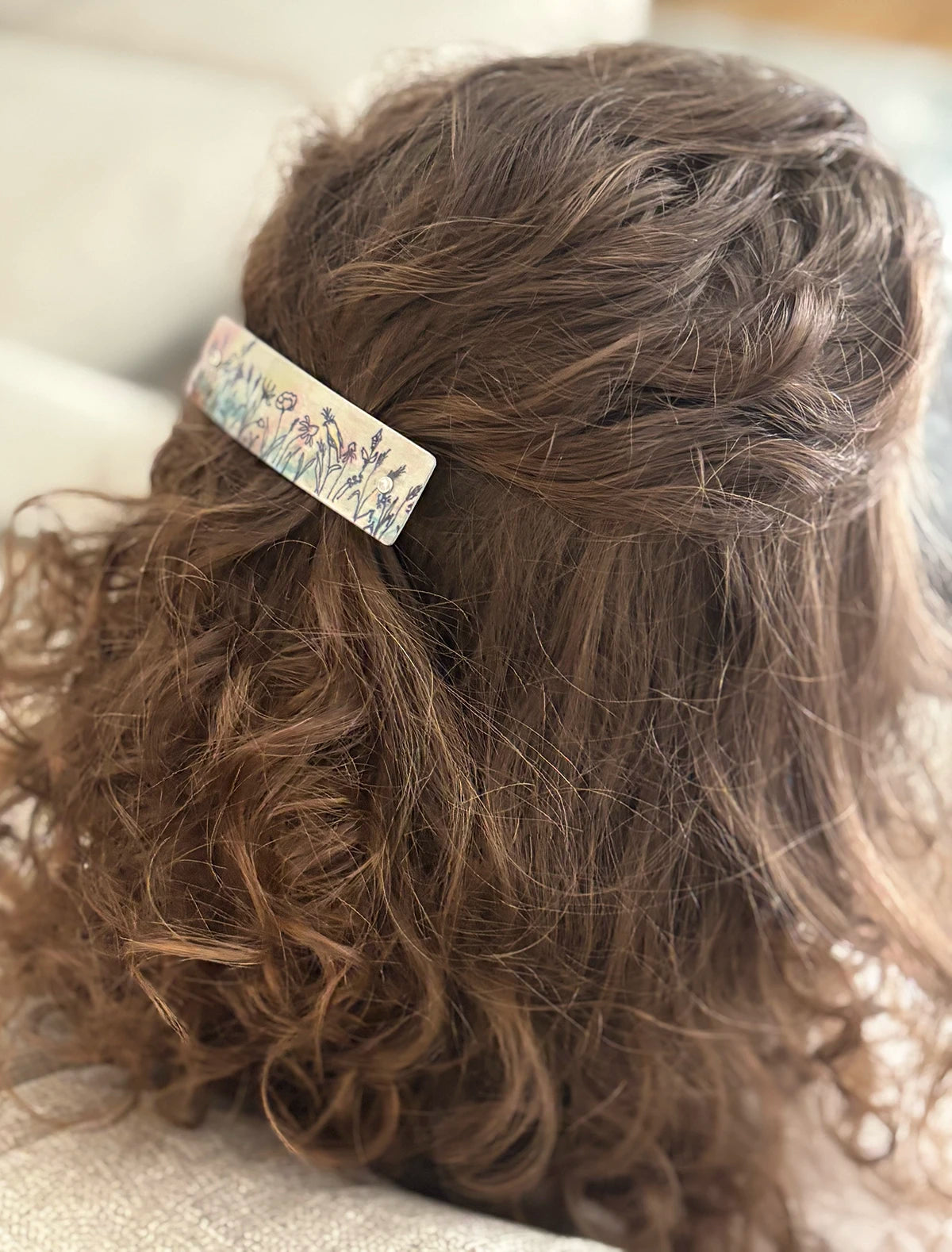 A close-up back view of a person with brown curly hair wearing a rectangular hair clip with a delicate bird and floral pattern on a light background.
