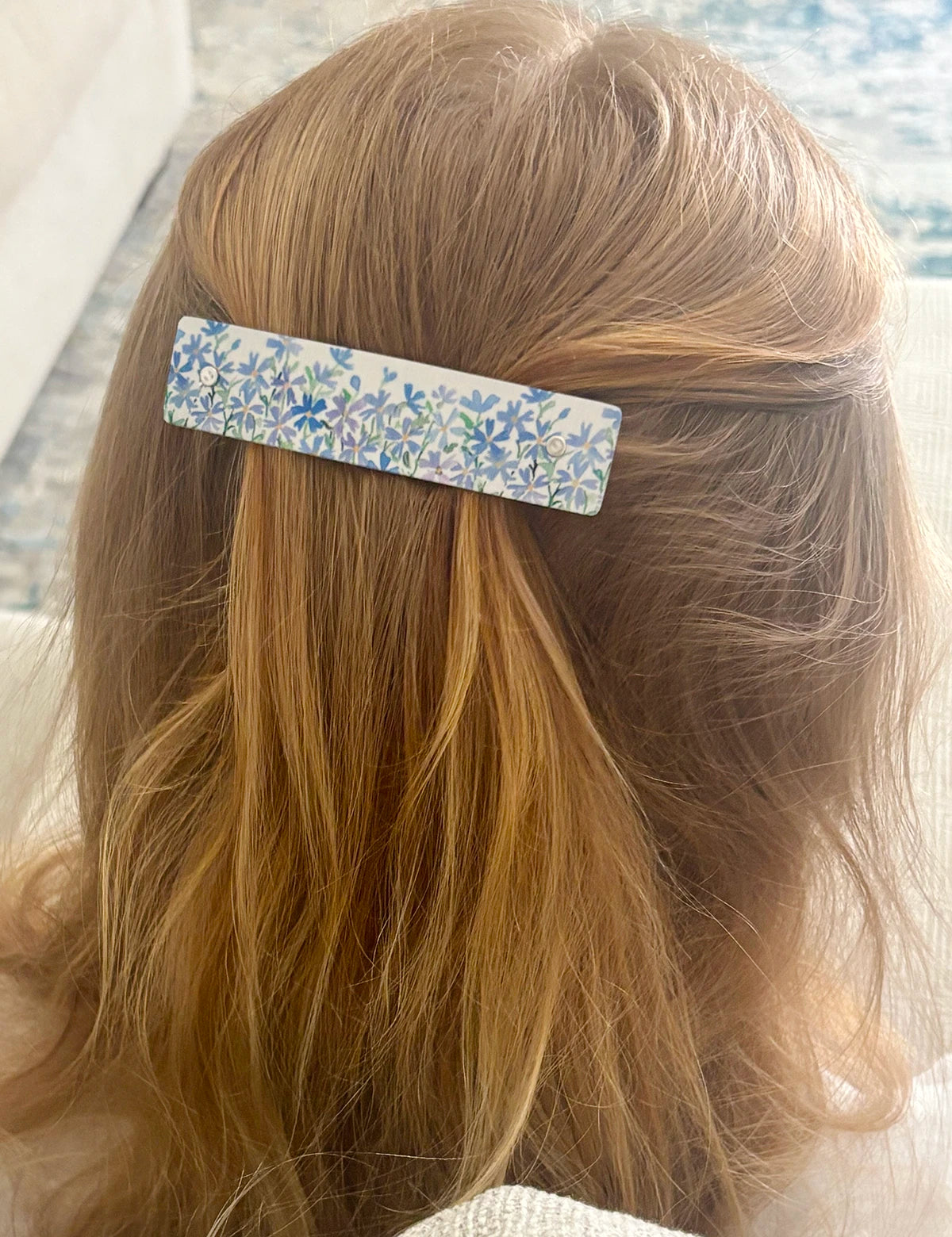 A close-up lifestyle shot of a person's blonde hair secured with a rectangular hair clip featuring a lavender and blue dainty floral pattern.