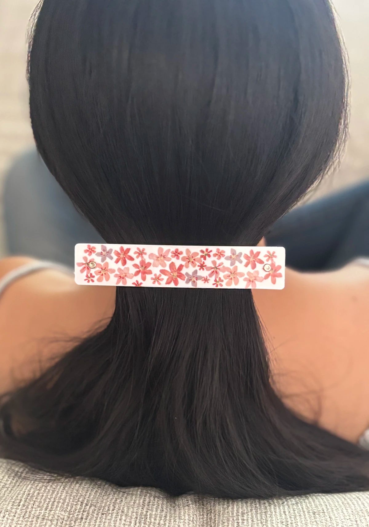 A close-up back view of a person with long black hair secured with a white rectangular hair clip decorated with a repeating red and pink floral pattern.
