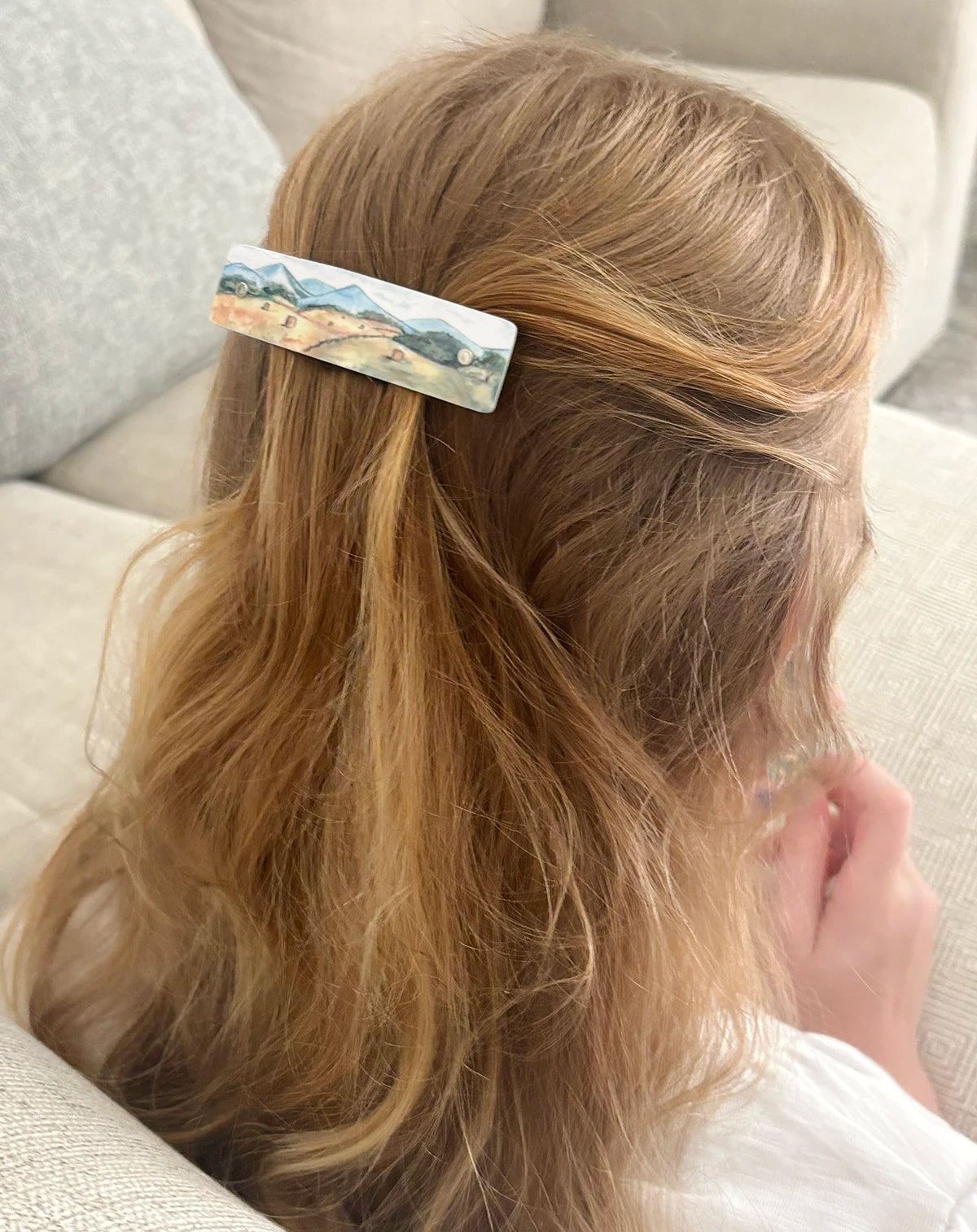 A woman with long blonde hair seen from behind, wearing a rectangular hair clip with artwork of a scenic landscape , seated on a light beige sofa.