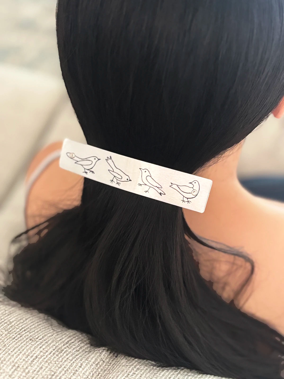 A close-up of a person's black hair secured with a silver rectangular hair clip decorated with funny, cute black bird illustrations.