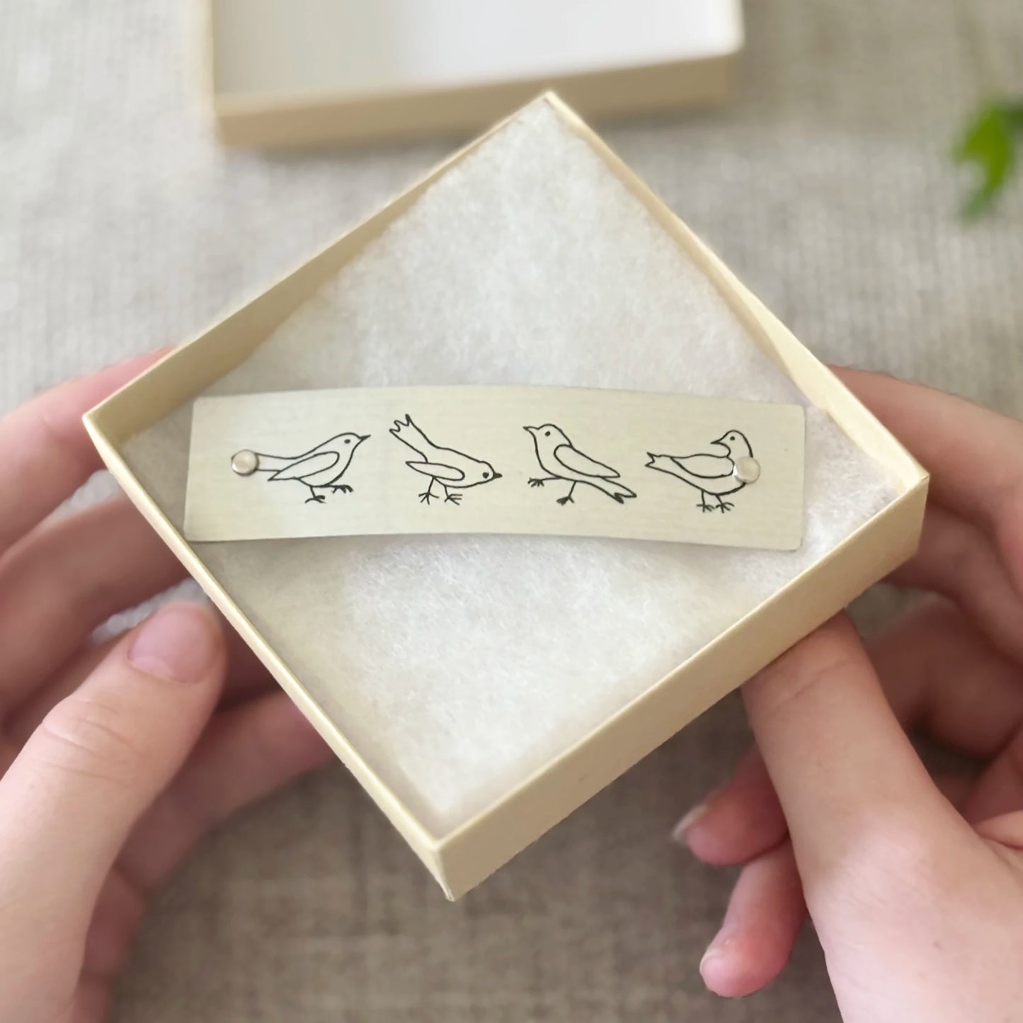 A beige rectangular hair clip with minimalist humorous black bird illustrations, held in a light beige gift box.
