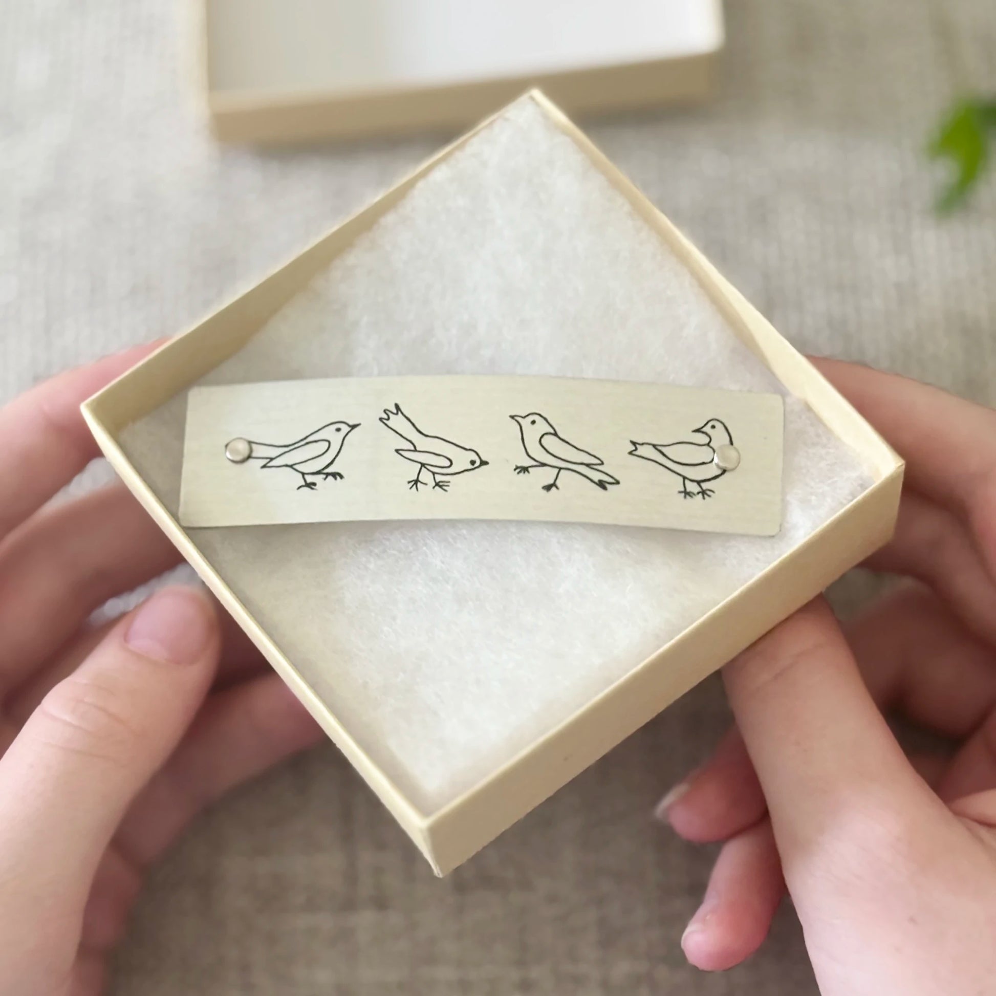 A beige rectangular hair clip with minimalist humorous black bird illustrations, held in a light beige gift box.