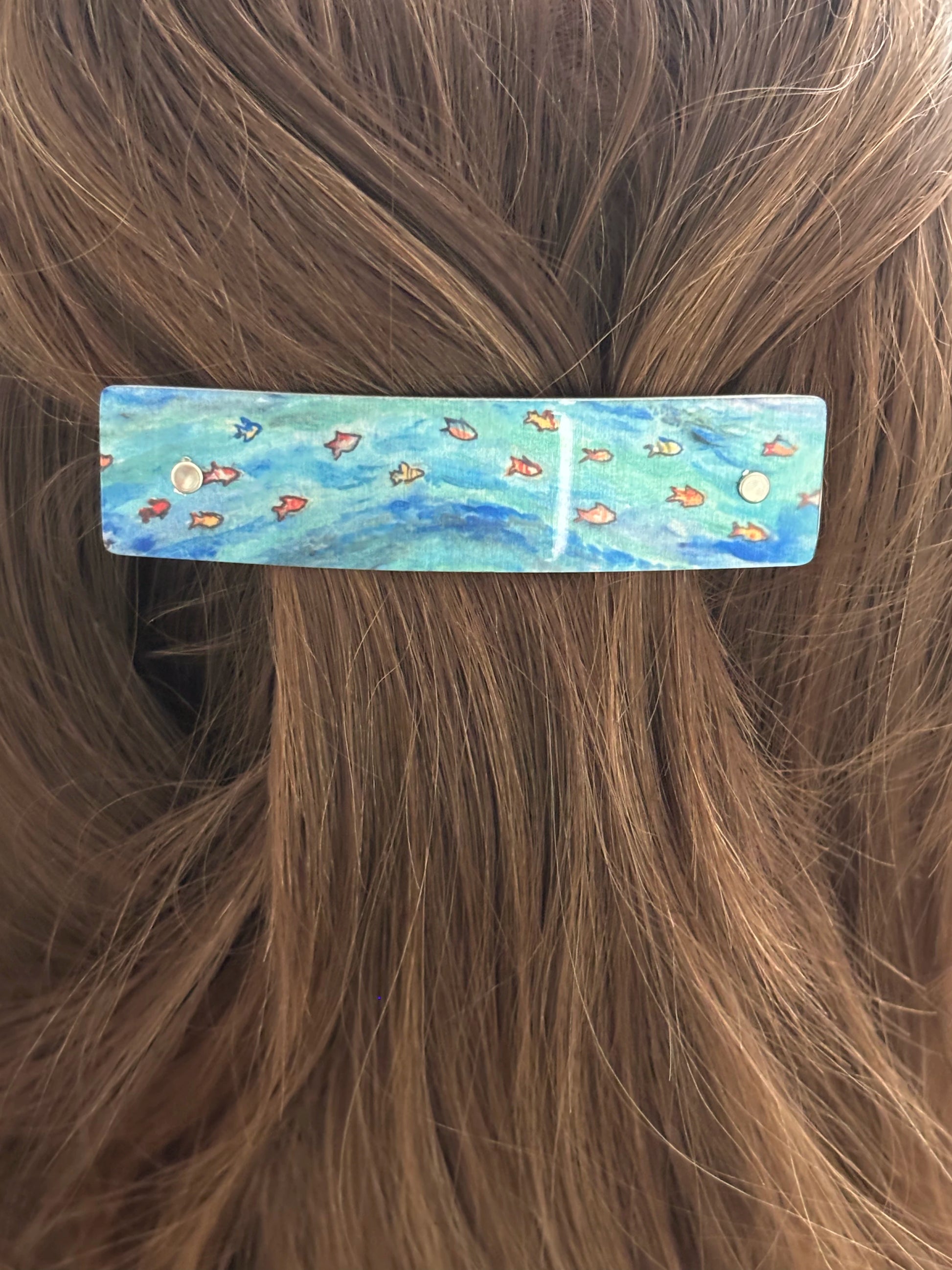 A rectangular hair barrette with a blue and green underwater fish pattern, shown clipped onto brown hair.