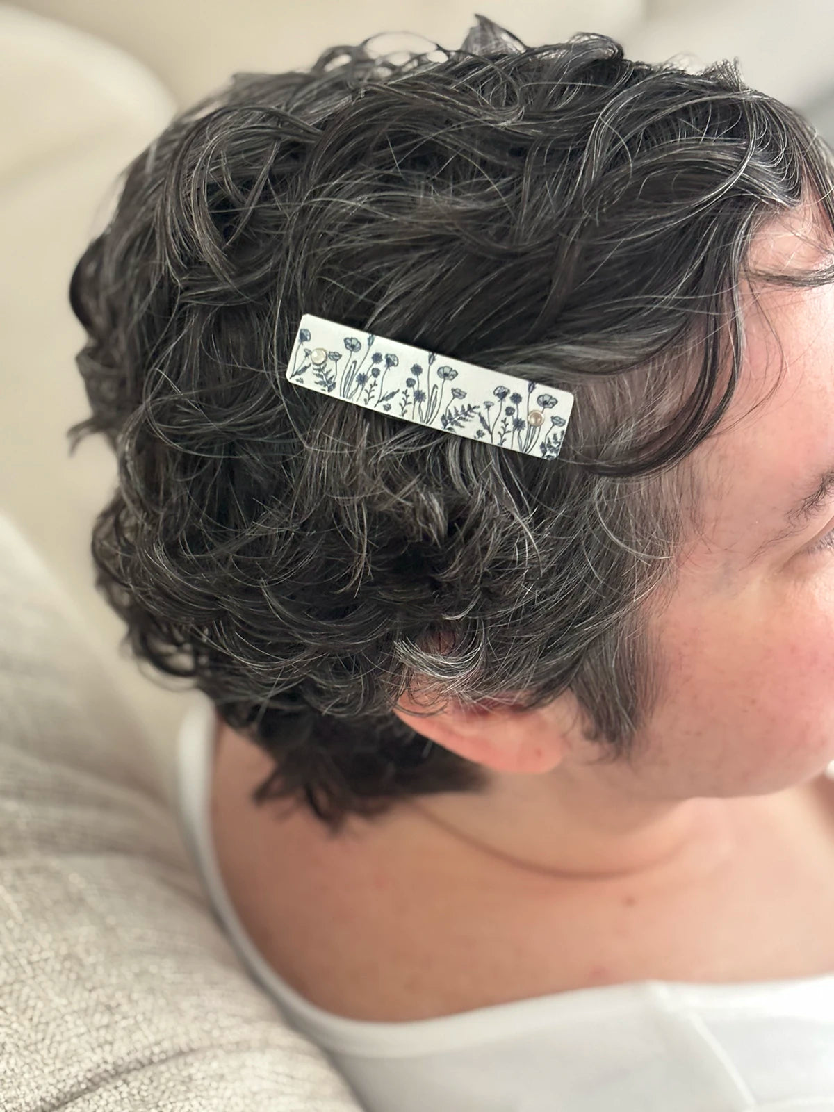 A close-up of a person with short, curly dark hair wearing a silver hair clip decorated with a black floral pattern.