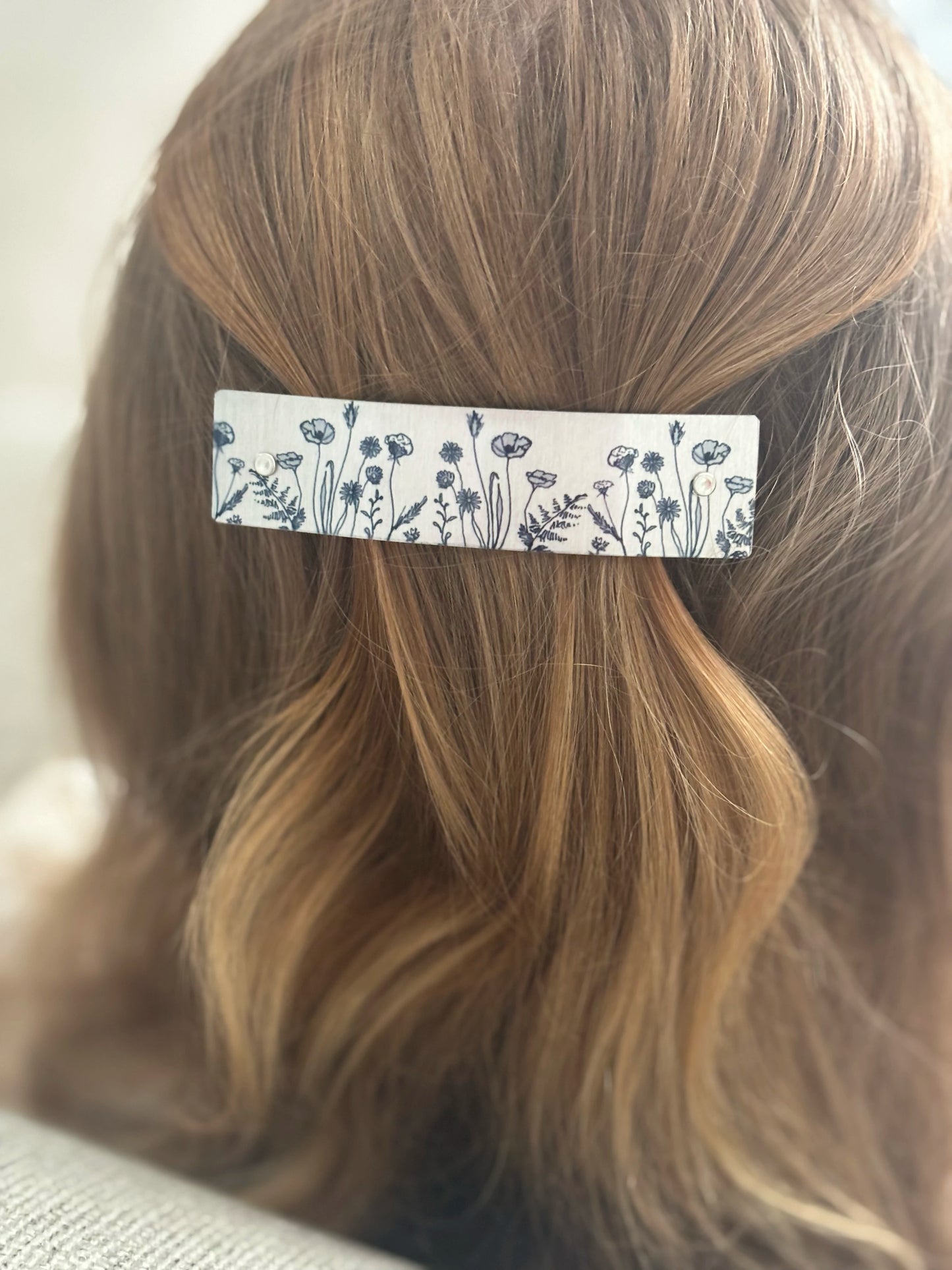 A close-up of a person's light brown hair secured with a rectangular hair clip featuring a black wildflower pattern on a silver background.
