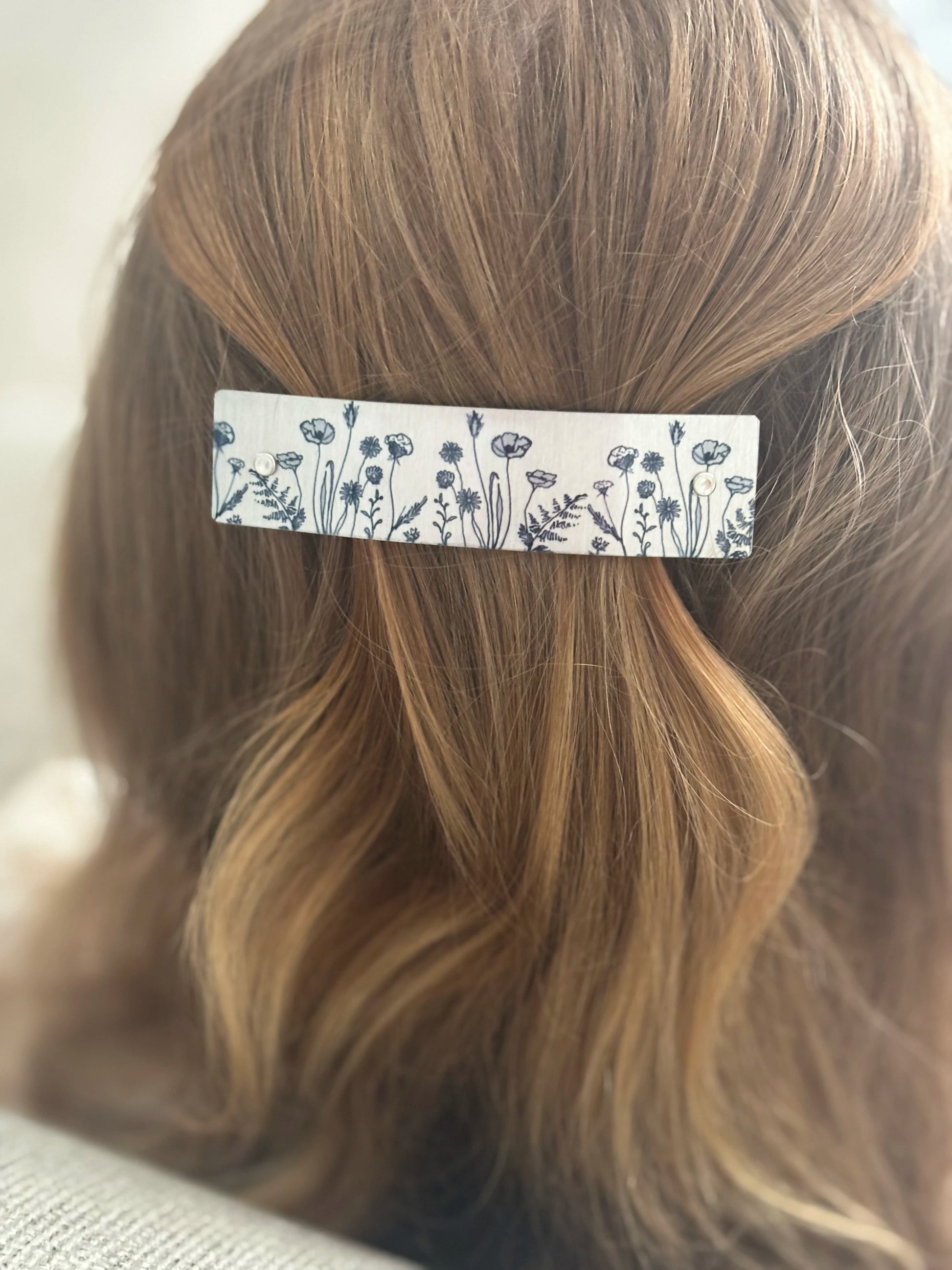 A close-up of a person's light brown hair secured with a rectangular hair clip featuring a black wildflower pattern on a silver background.