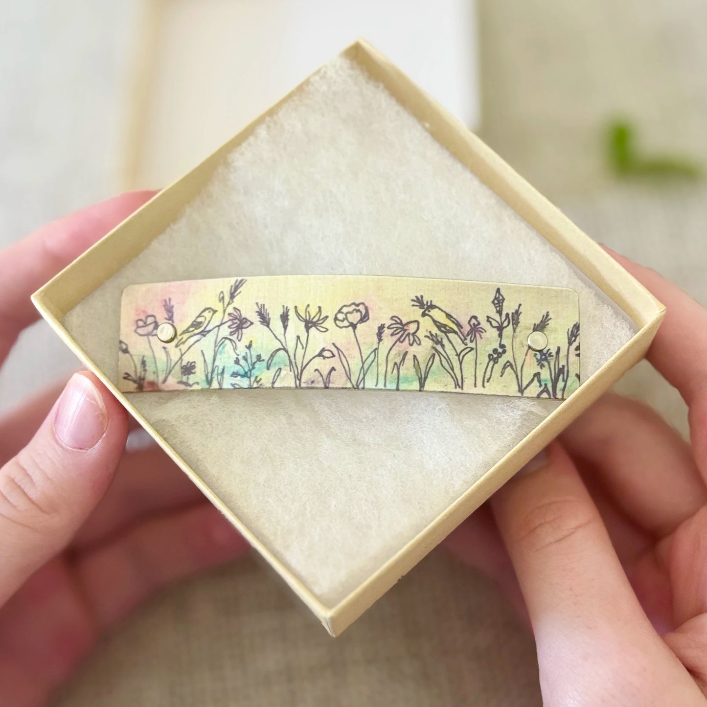 A close-up of a hand holding a small rectangular box with a floral and bird patterned barrette inside, ready for gifting.