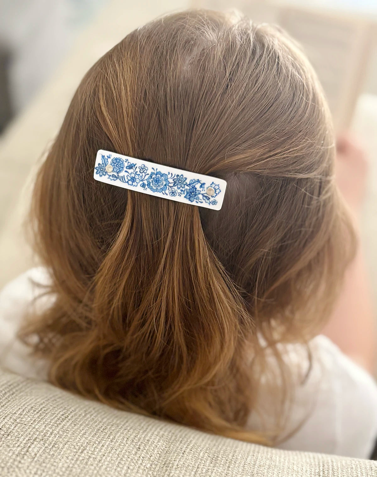 A close-up back view of a person with light brown hair wearing a white hair clip decorated with a blue floral Delft pattern.