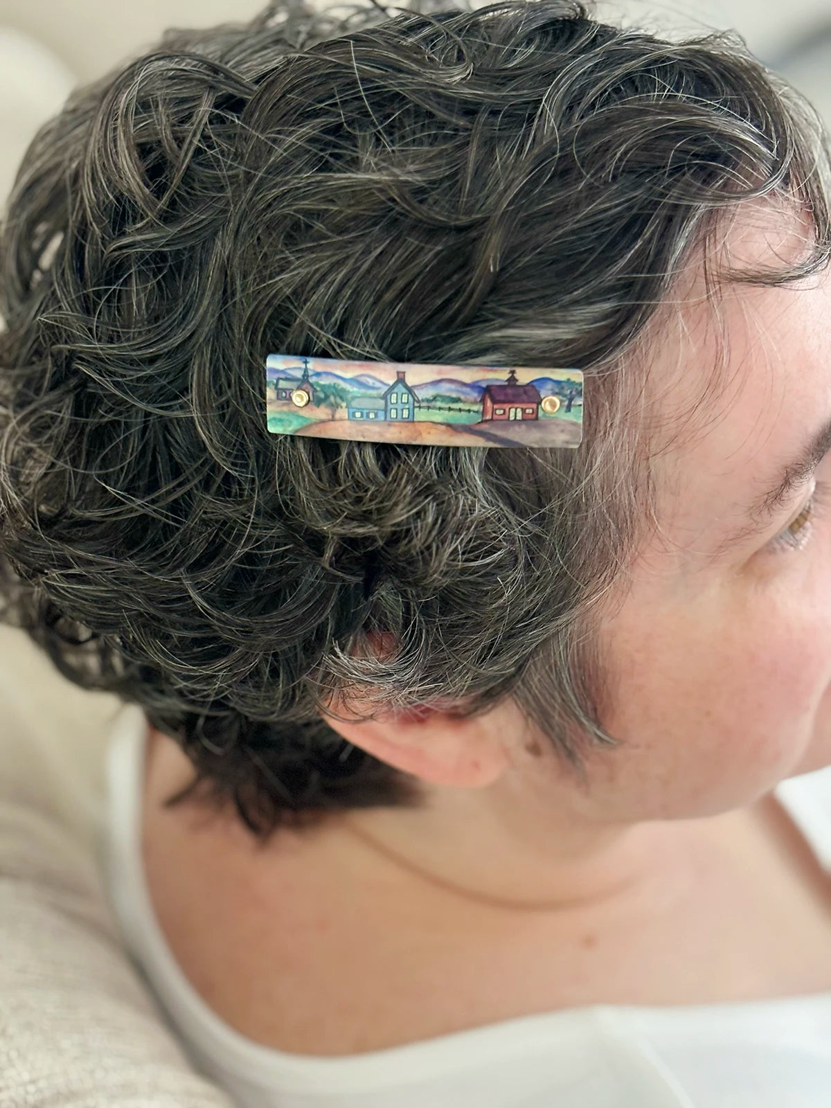 A close-up side view of a person with short, curly gray hair wearing a small rectangular hair clip decorated with a colorful landscape illustration.