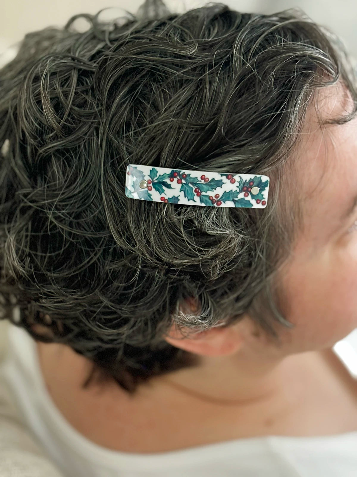 A close-up of a person's short, curly dark gray hair wearing a rectangular hair clip decorated with a green holly and red berries pattern on a white background.