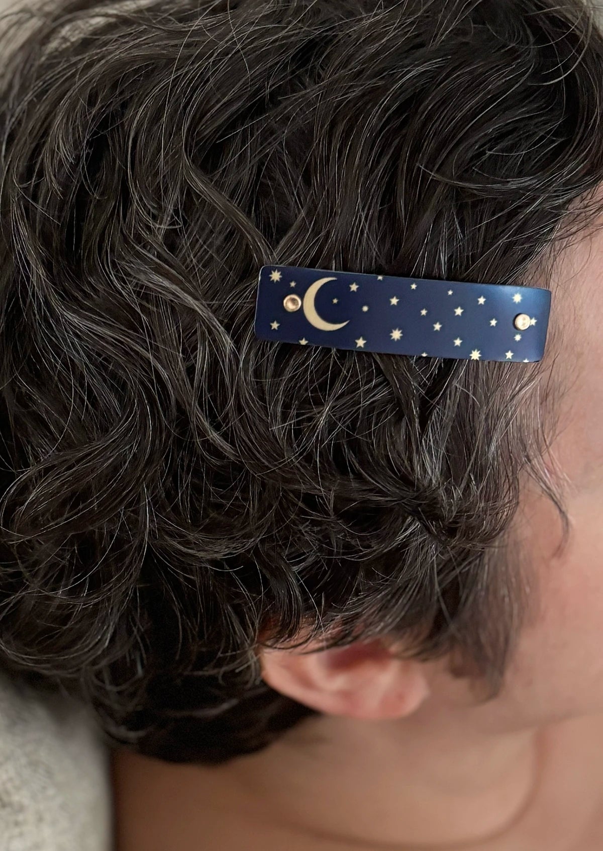 A close-up of a person's dark curly hair wearing a navy blue hair clip decorated with gold celestial patterns of stars and a crescent moon.