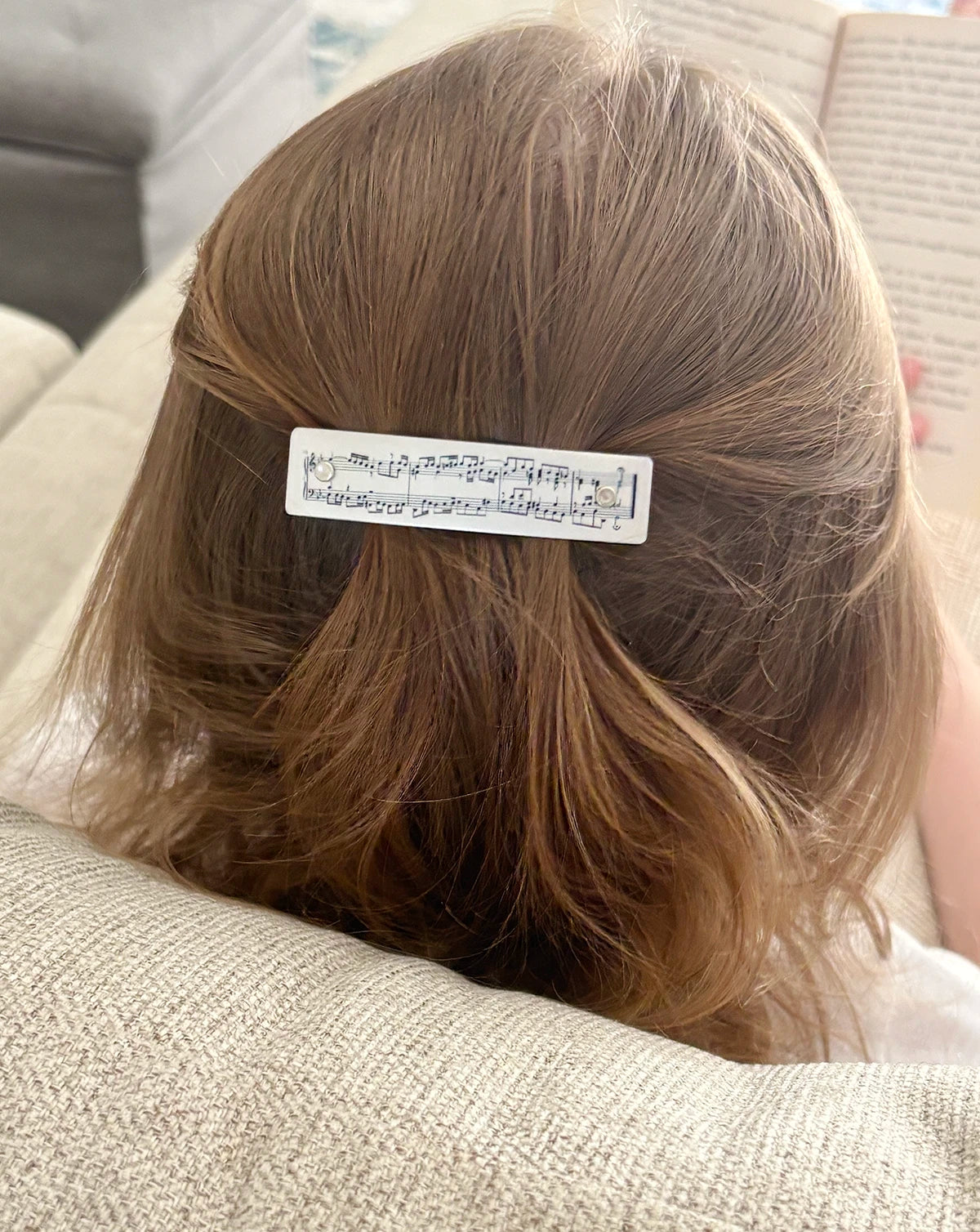 A close-up lifestyle shot of a child's brown hair from behind, featuring a silver hair clip decorated with black musical notation, resting on a beige textured couch next to an open book.
