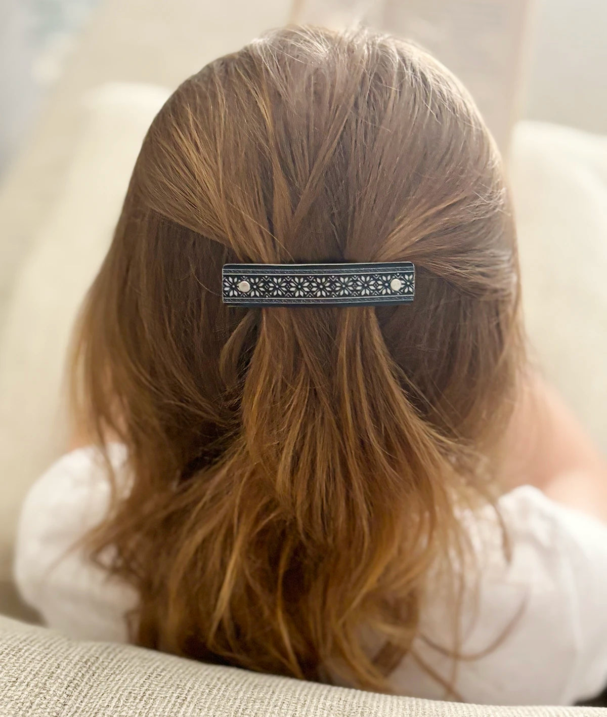 A close-up back view of a child with light brown hair wearing a slim black hair clip adorned with small silver floral details, resting on a light beige sofa.