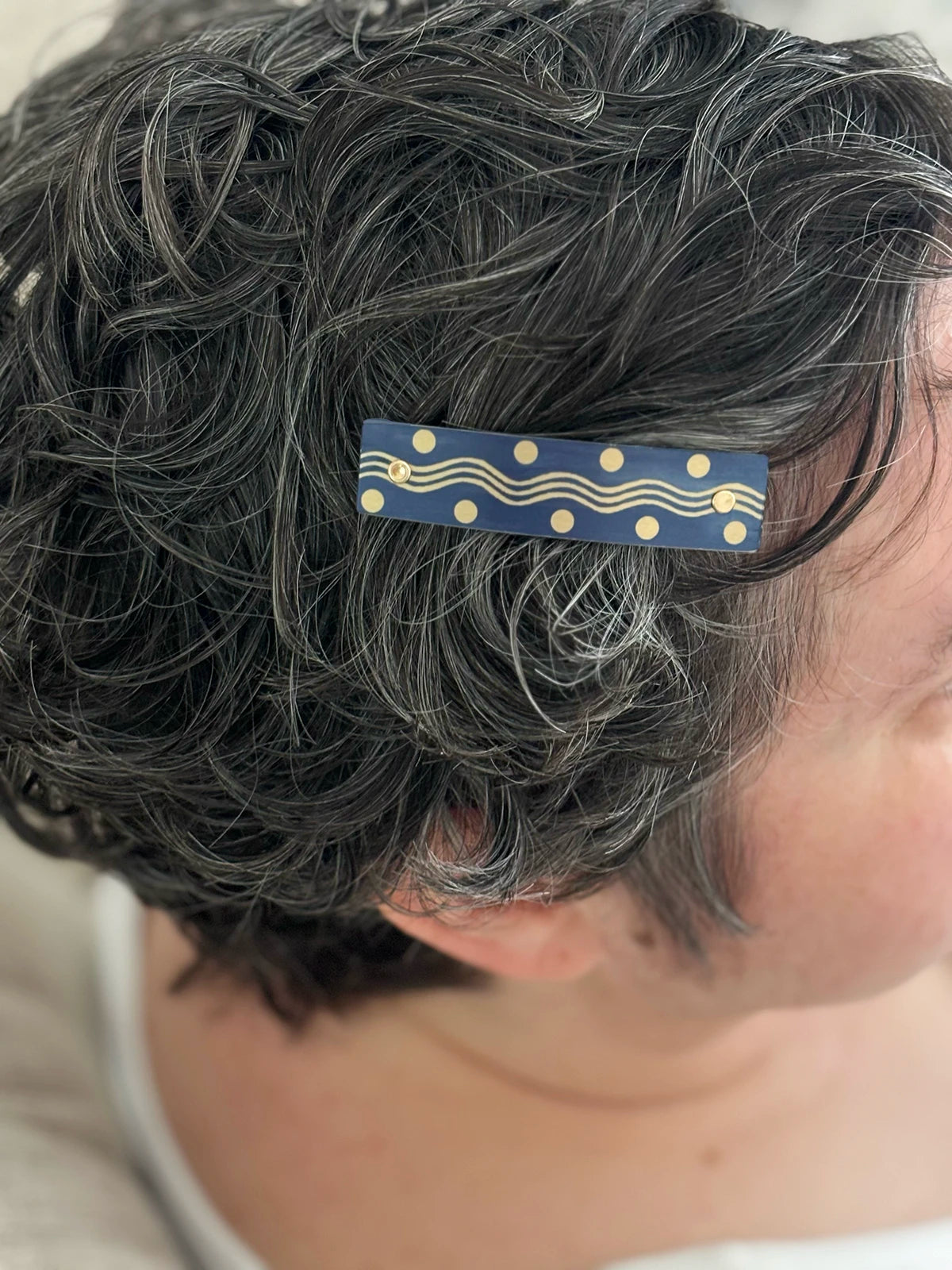 A close-up of a person's short, curly gray hair wearing a navy blue-gray hair clip with gold wavy lines and dots.