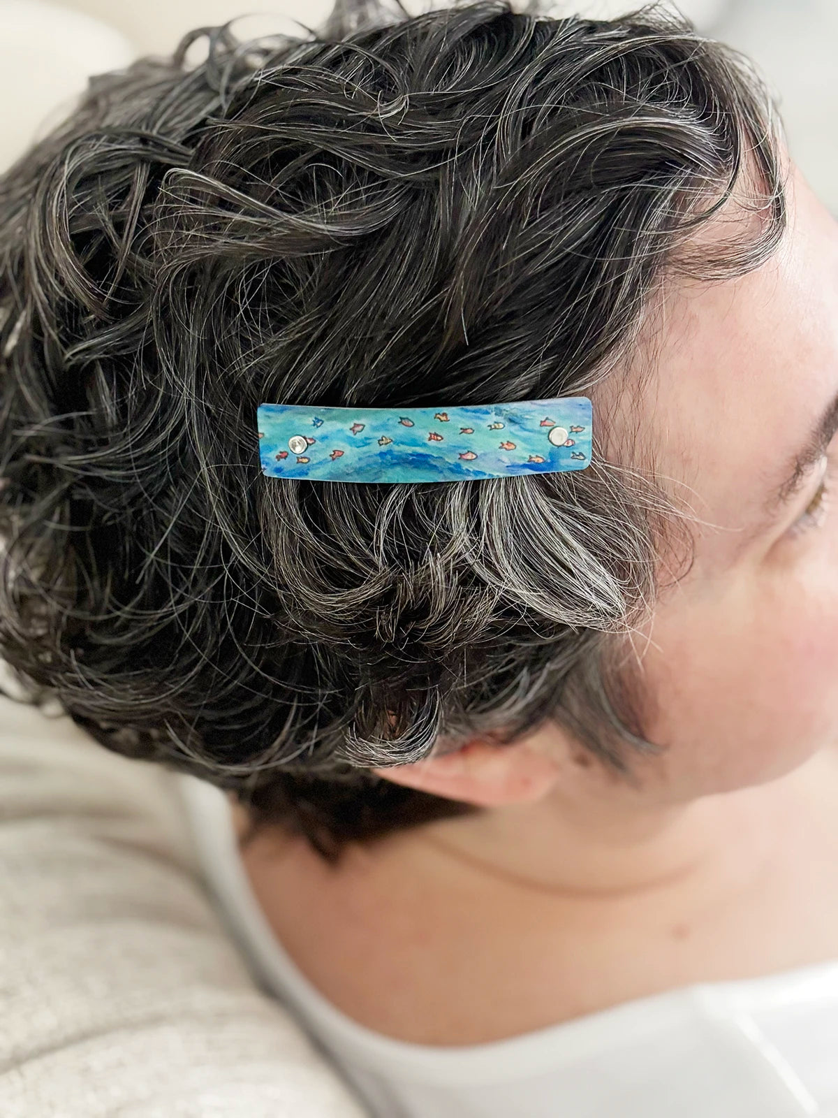 A close-up of a person with short, curly gray hair wearing a rectangular hair clip. The clip is light blue and green with a pattern of tiny tropical fish.