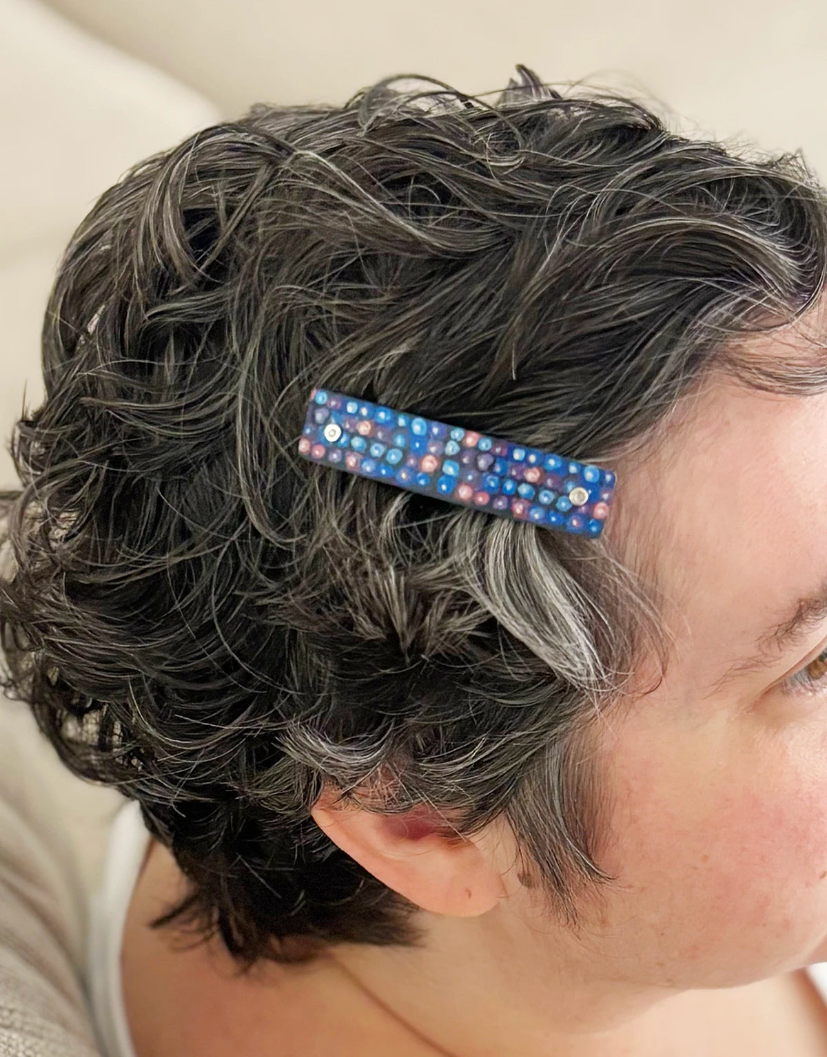 A close-up side view of a person with short, curly gray hair wearing a decorative hair clip. The hair clip is rectangular and features a blackberry inspired design.