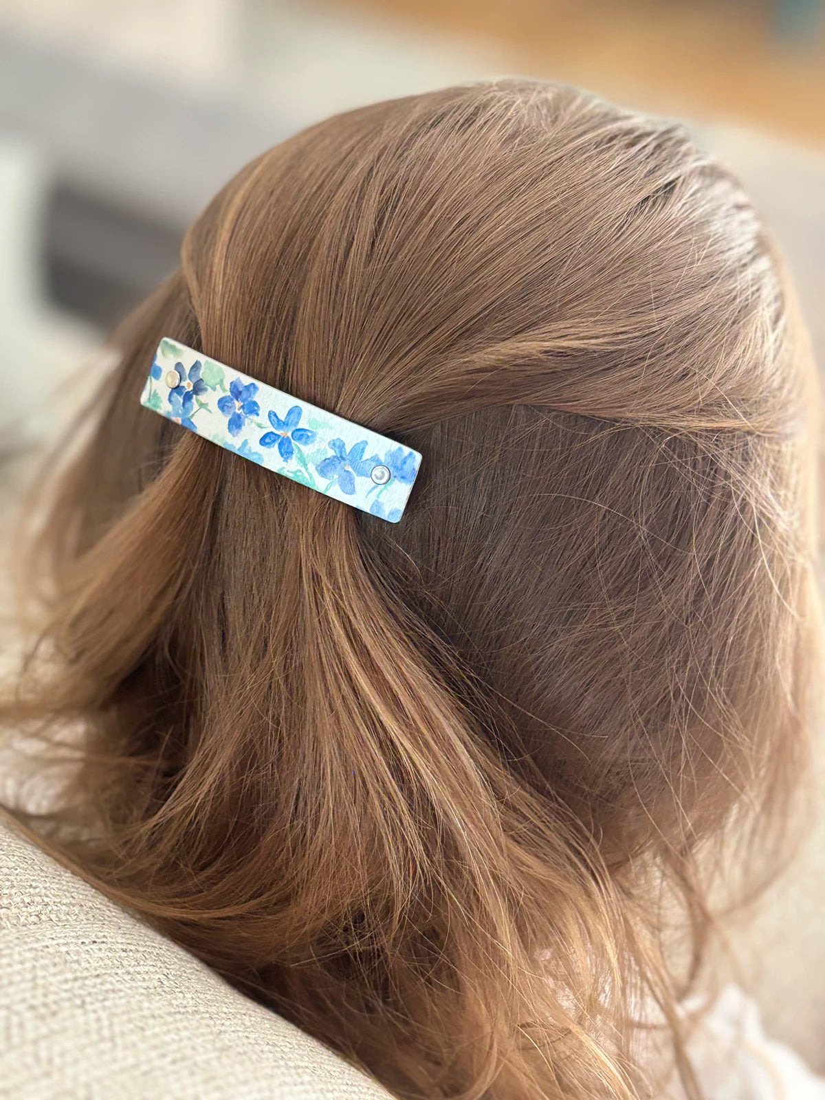 A close-up of a child's light brown hair secured with a slim rectangular hair clip featuring a blue floral pattern on a silver background.