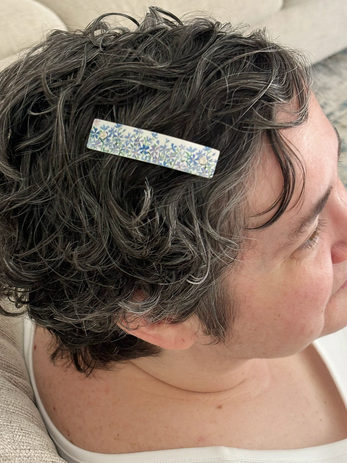 A close-up side profile of a person with short, curly gray hair wearing a rectangular hair clip with a blue and purple floral phlox pattern.