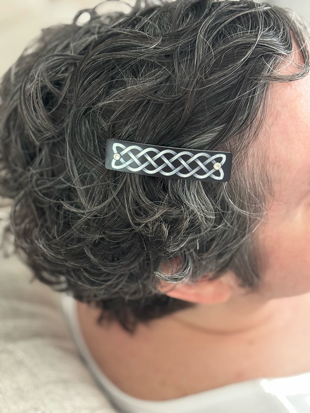 A close-up of a person's dark gray and silver curly hair, featuring a black hair clip with a silver Celtic knot design.