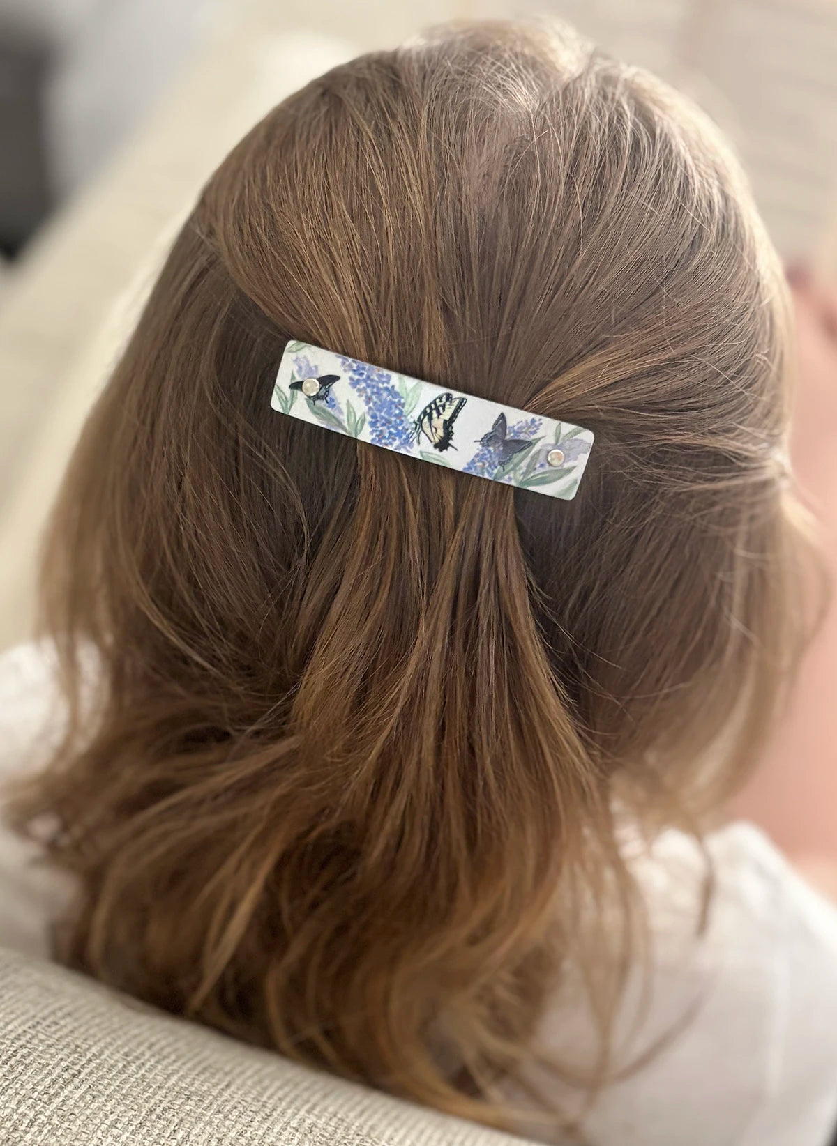 A close-up back view of a child with light brown hair wearing a slim rectangular hair clip with a blue and silver floral pattern.