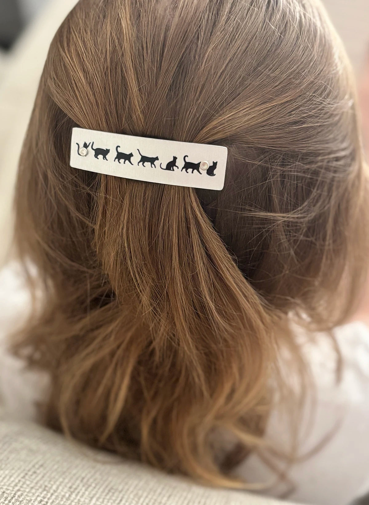 A close-up of a person's light brown hair from behind, featuring a silver rectangular hair clip decorated with black cat silhouettes, held in place.