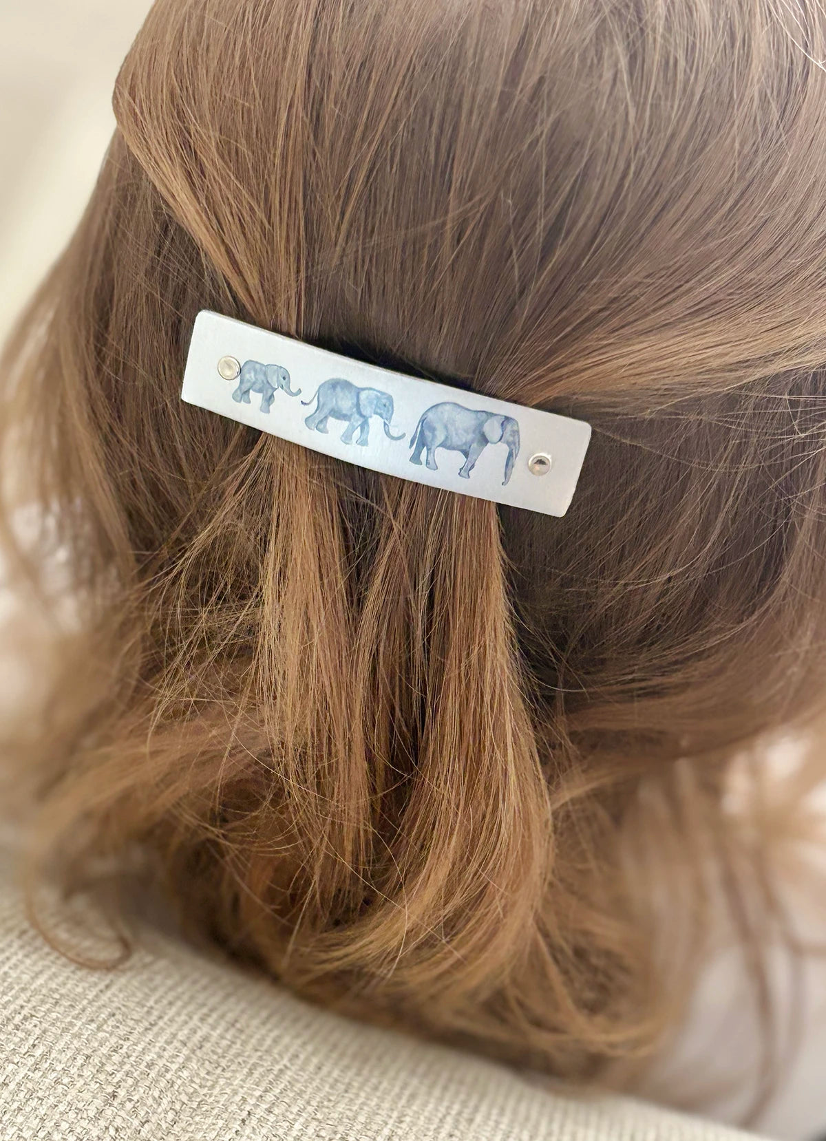 A close-up of a child's light brown hair secured with a metal barrette hair clip decorated with watercolor elephant illustrations.