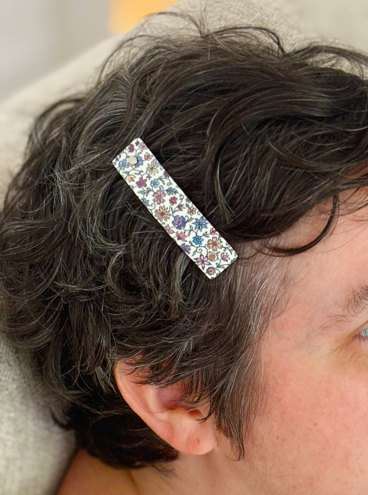A close-up of a person's dark brown hair wearing a slim rectangular hair clip decorated with a delicate multicolor floral pattern on a white background.