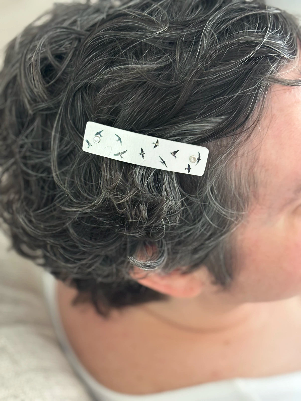 A close-up of a person's short, curly gray hair with a silver rectangular hair clip featuring small black bird silhouettes, shown from the side.