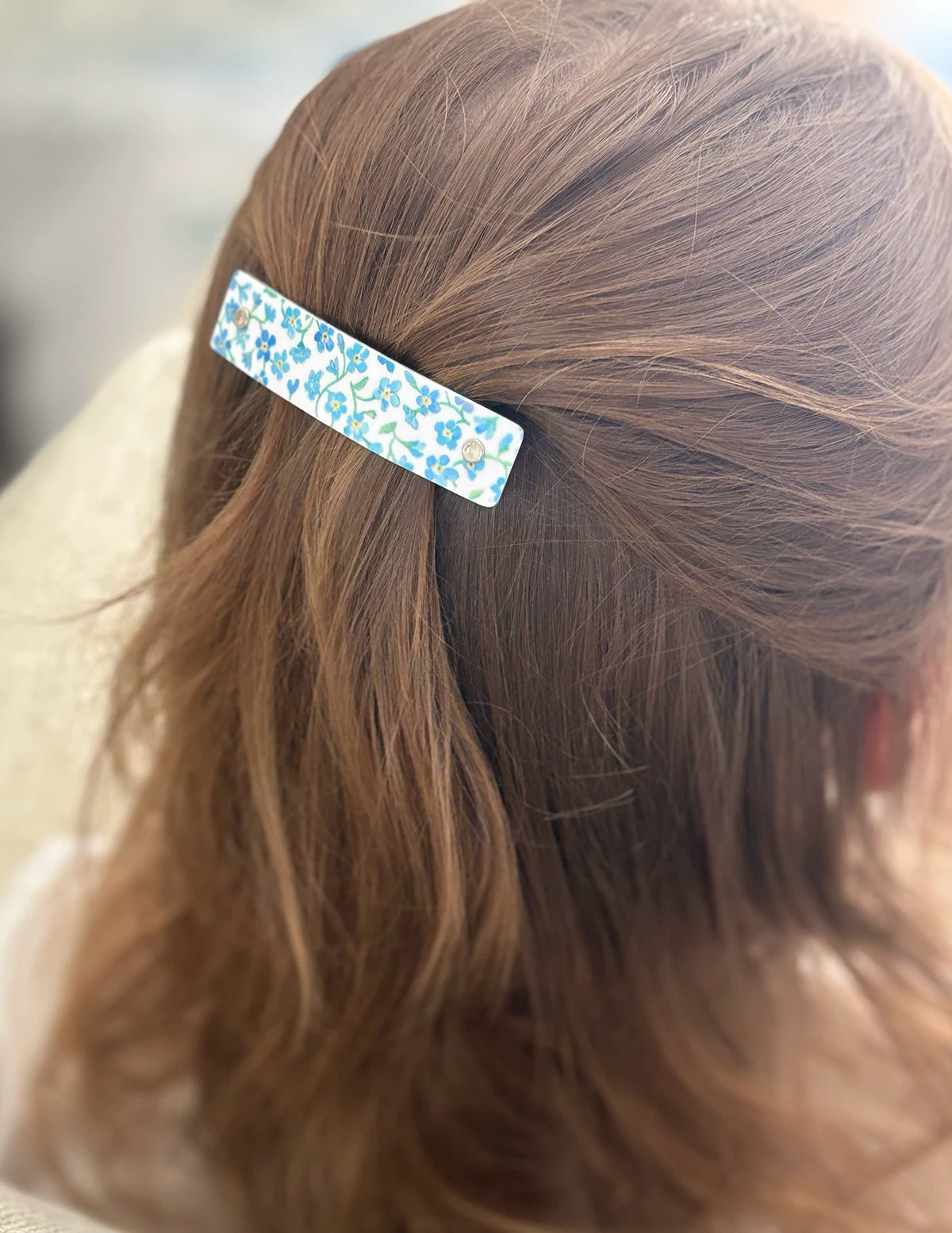 A close-up shot of a child's brown hair secured with a small rectangular barrette hair clip featuring a light blue and white floral pattern.