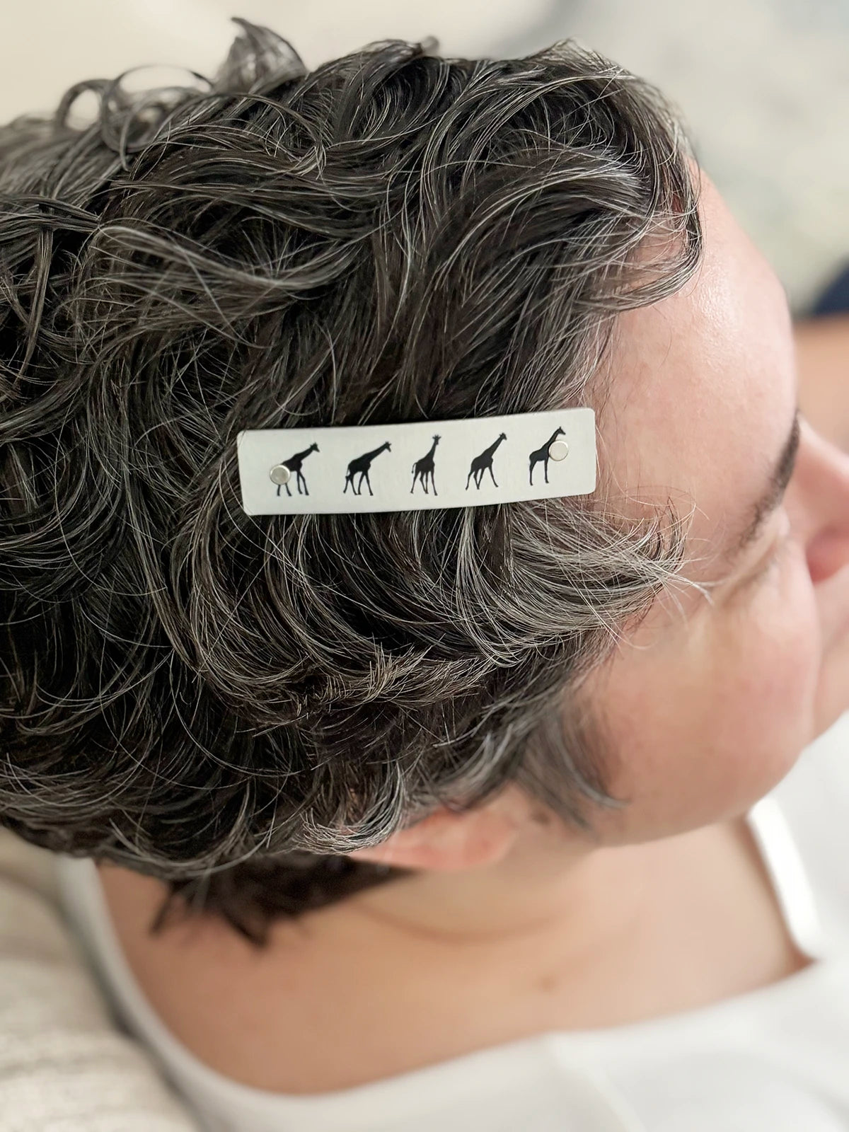 A close-up of a person's short, curly gray hair with a silver hair clip featuring black giraffe silhouettes.