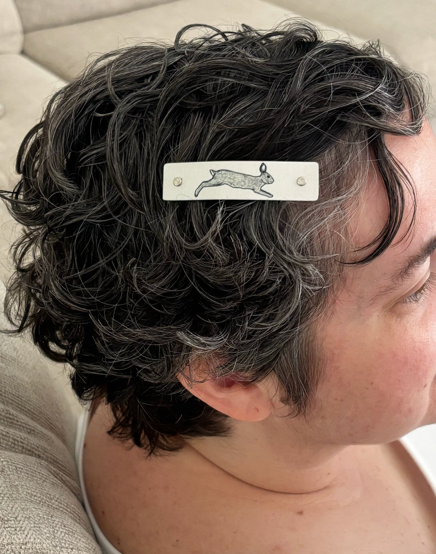 A close-up side view of a person with short, curly gray hair wearing a silver hair clip with a gray rabbit drawing..