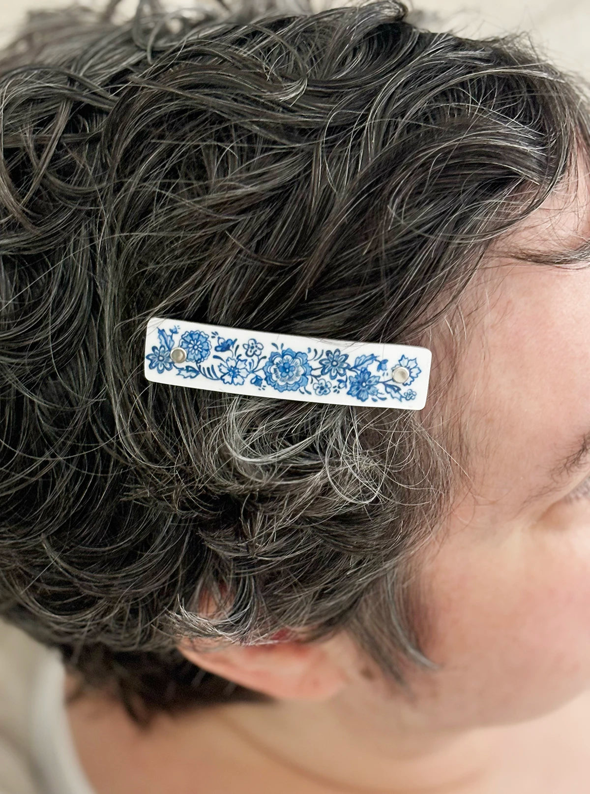 A close-up of a person's dark, curly hair with a white hair clip featuring a traditional blue floral pattern, worn on the side of the head.
